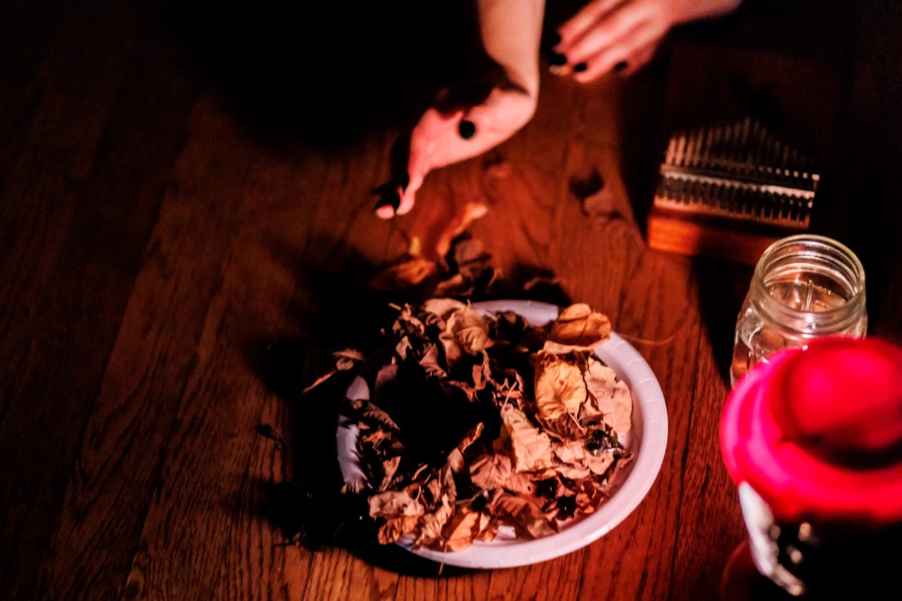 A person with black nail polish arranges dried leaves on a plate, next to a glass jar and a red candle, on a wooden surface in dim lighting.