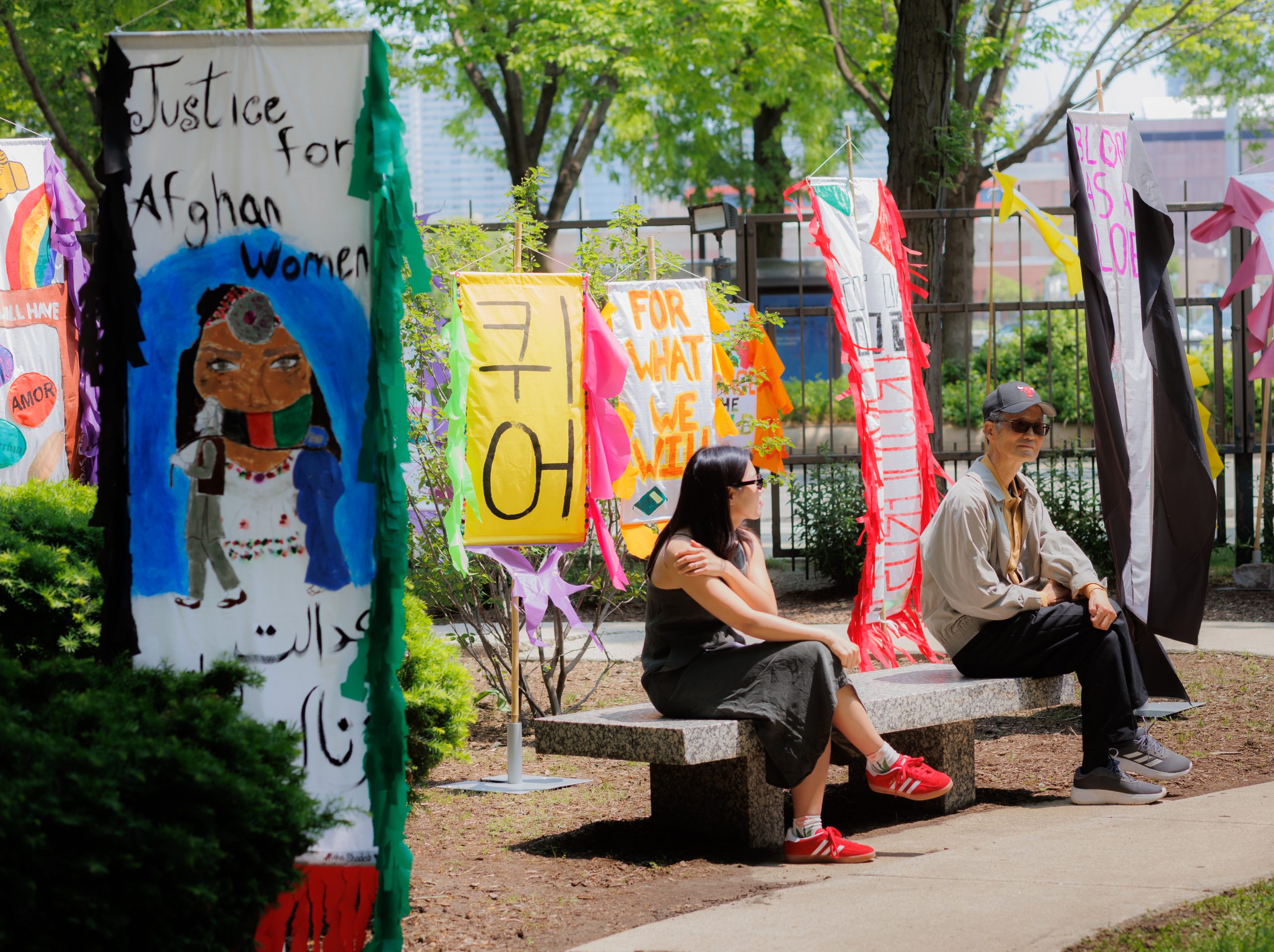 Two people sit on a bench in a park surrounded by colorful banners and signs, including one that reads Justice for Afghan Women. Trees and greenery are visible in the background.