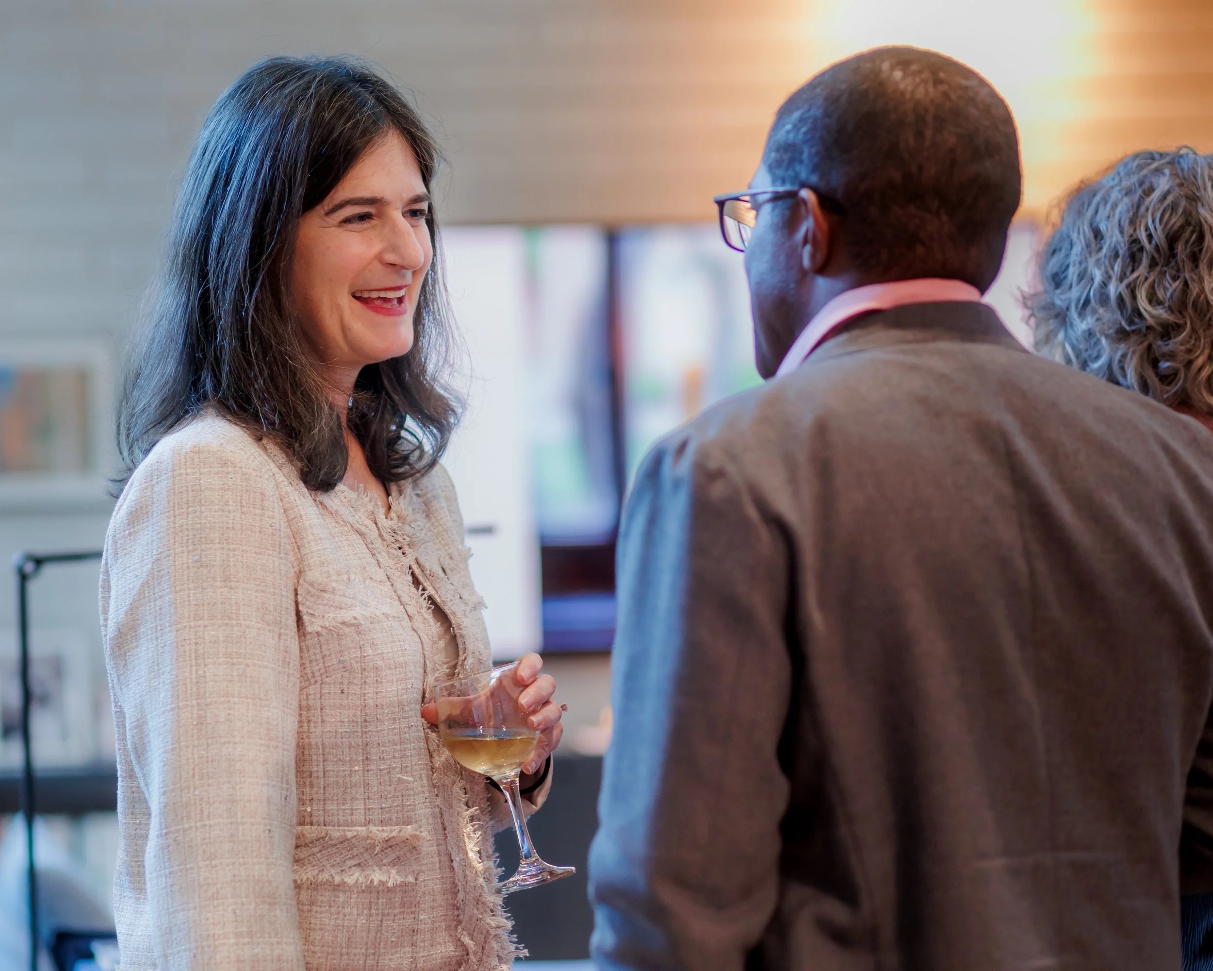 A woman holding a drink smiles while talking to a man in a suit at an indoor social event. Another person, partially visible, stands nearby. The atmosphere appears relaxed and friendly.