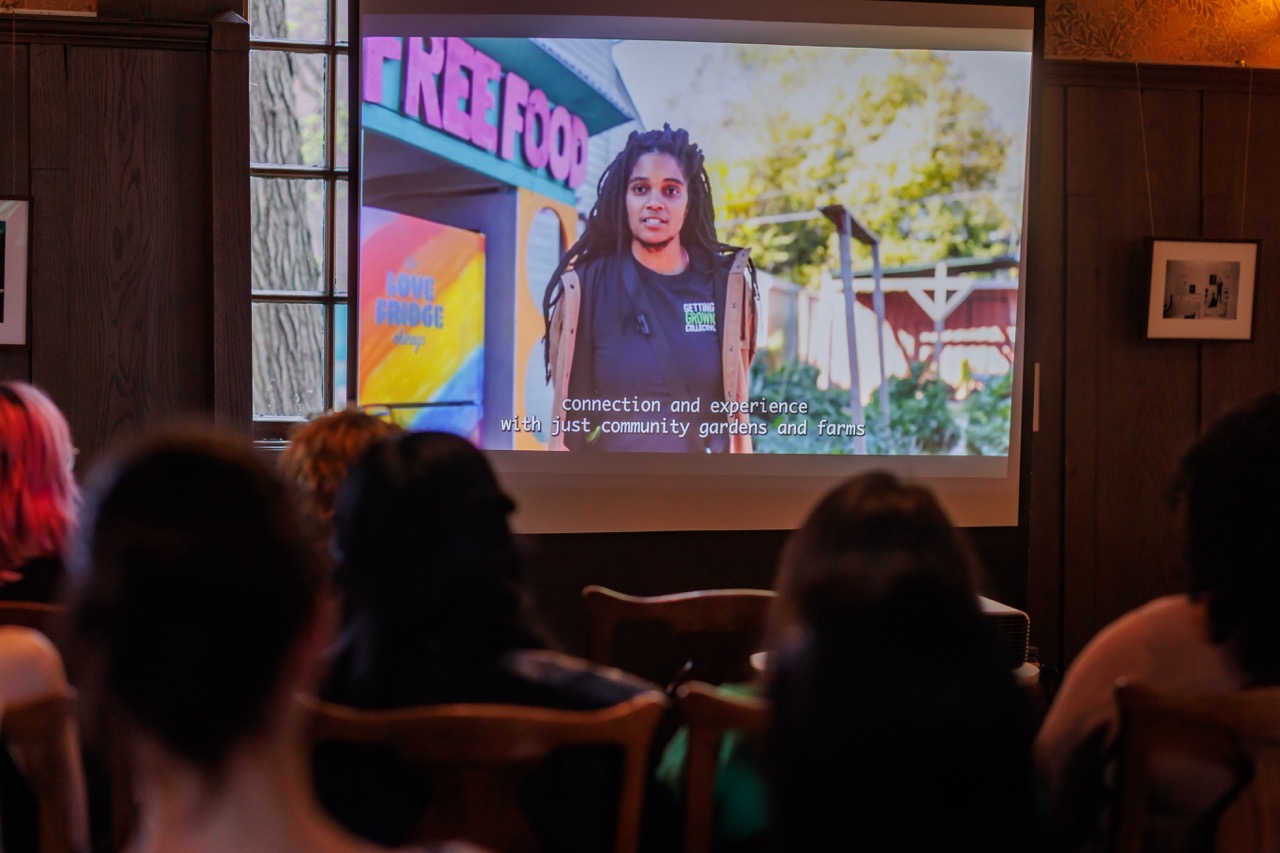 A group of people watch a film in a dimly lit room. The screen shows a woman speaking outside near a colorful structure labeled FREE FOOD. Subtitles are visible at the bottom of the screen.