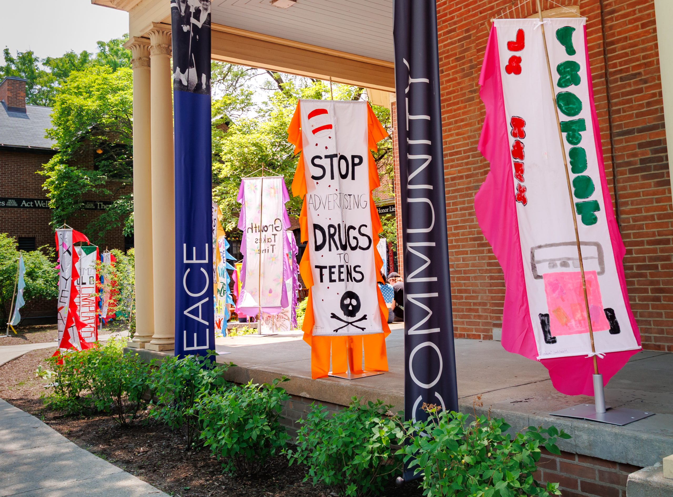 Colorful banners with anti-drug messages and drawings hang from columns outside a brick building. Two large banners read “PEACE” and “COMMUNITY.” Lush green plants line the walkway under the banners.