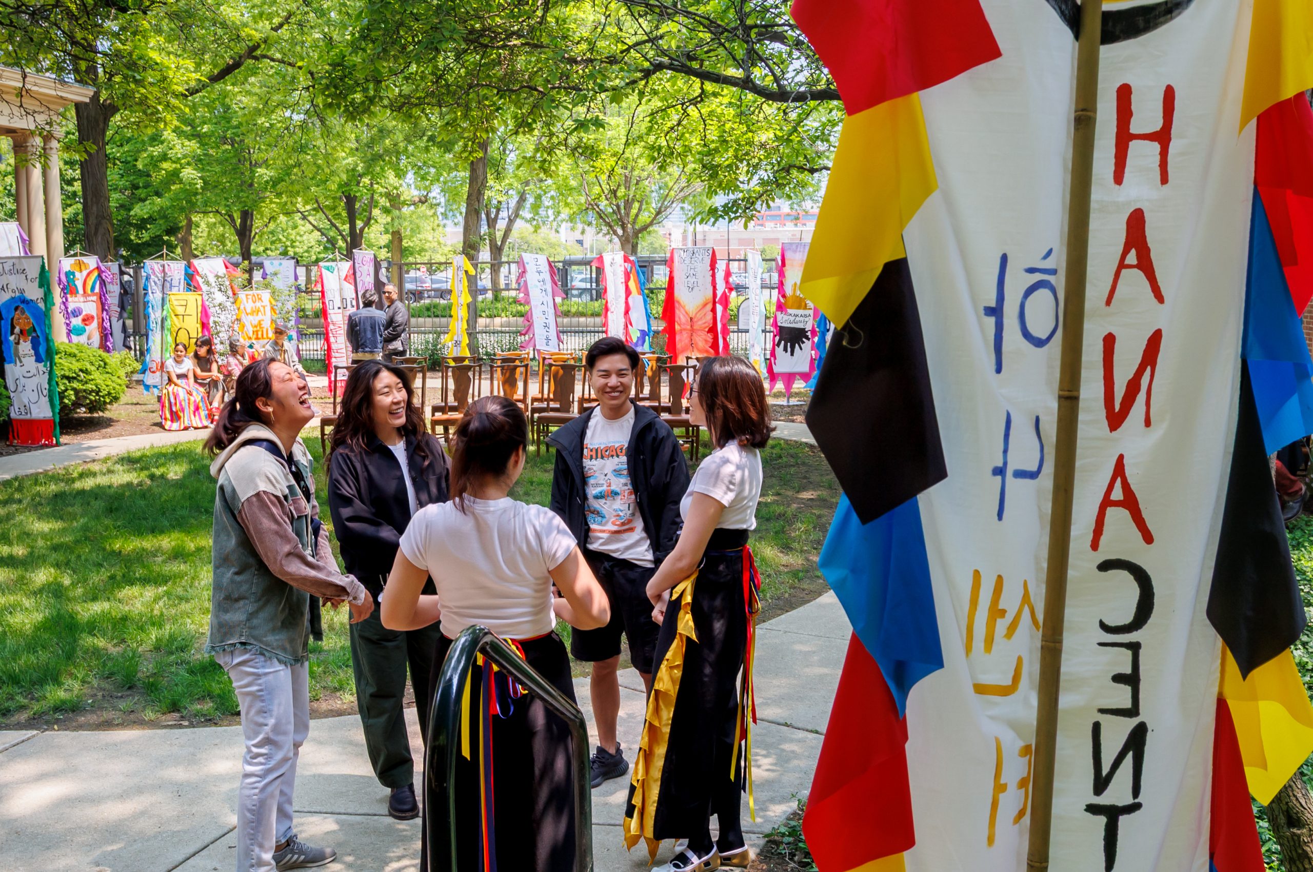 A group of five people stand in a circle, smiling and talking at an outdoor event decorated with colorful banners and flags featuring various designs and languages, surrounded by trees and greenery.