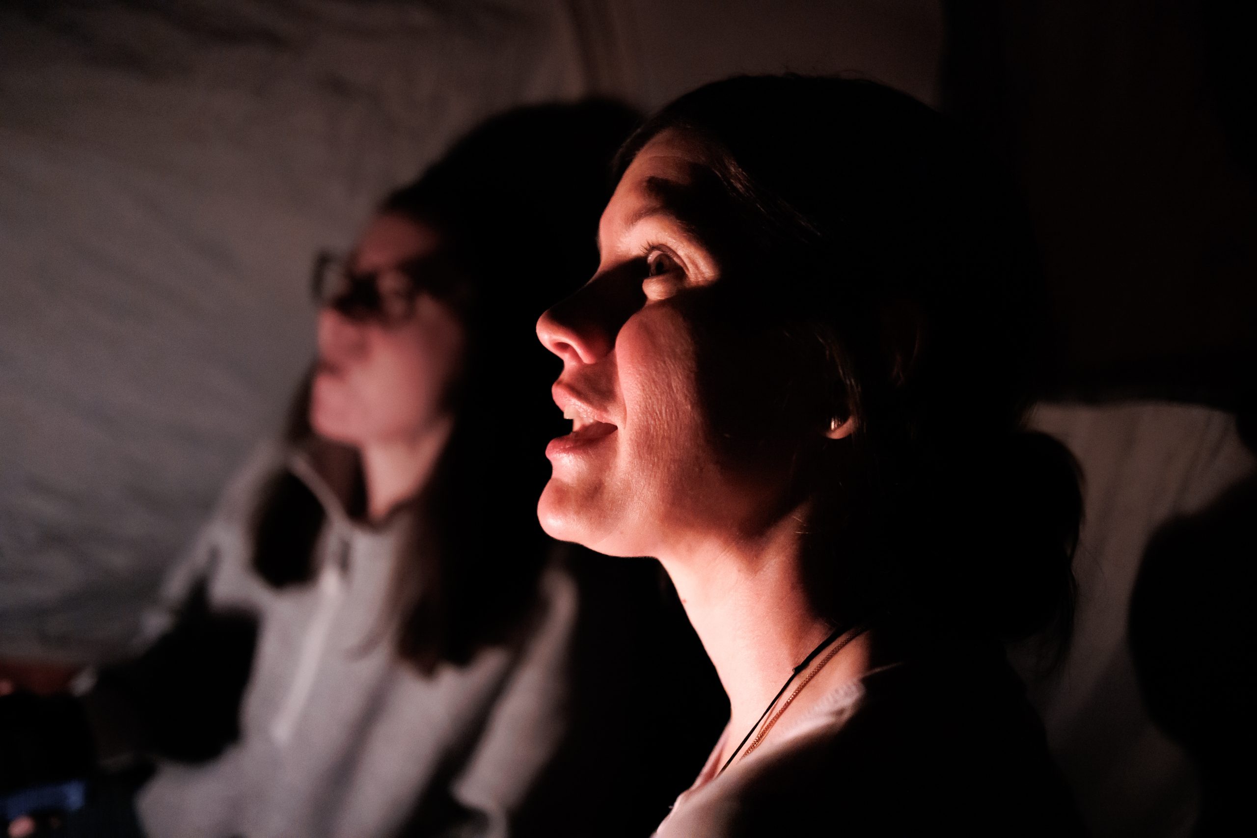 Two women sit in dim lighting; the woman in the foreground is illuminated and looking upward, while the woman in the background is blurred and partially in shadow.