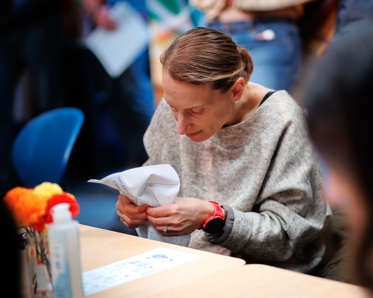 A woman with light brown hair in a low bun and wearing a grey sweater is carefully folding a piece of paper while sitting at a table. Colorful craft materials and a bottle are visible on the table.