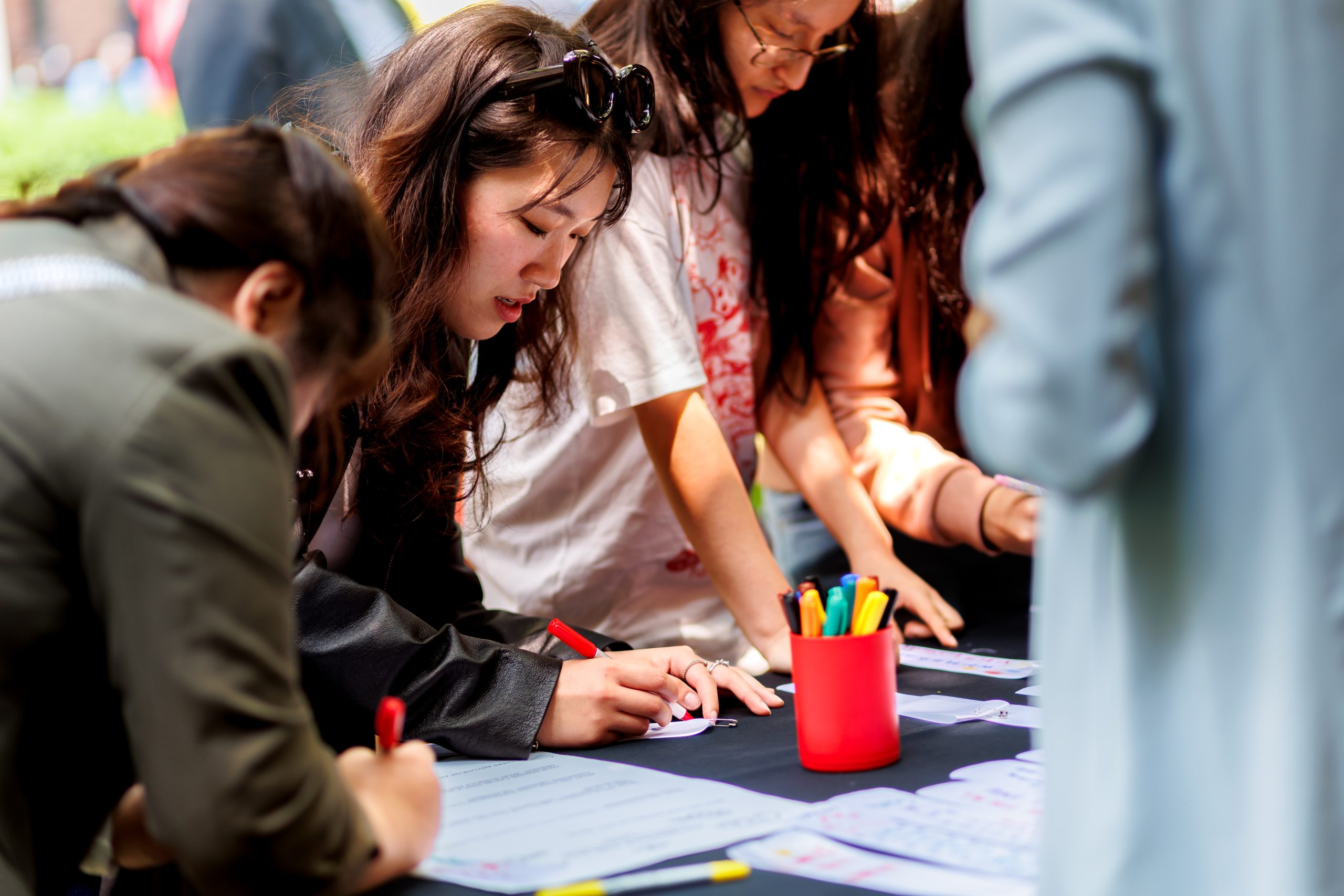 Several young people are gathered around a table, writing or drawing on papers with colorful markers. A red cup filled with markers sits on the table. The setting appears to be outdoors.