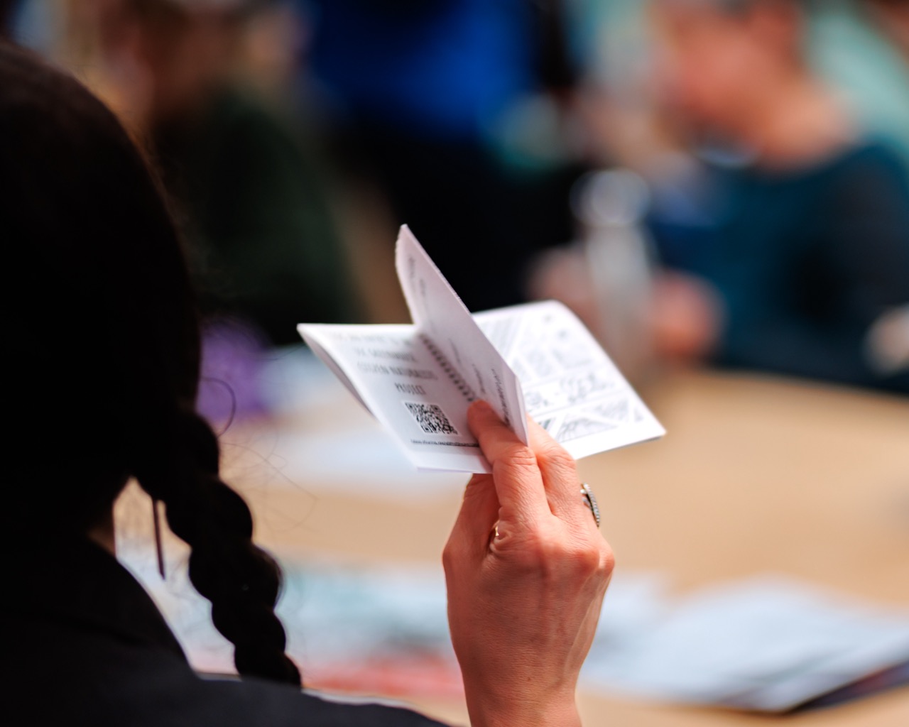 A person with a braid holds paper documents, possibly tickets or forms, while seated at a table. The background is blurred, showing other people sitting and working.