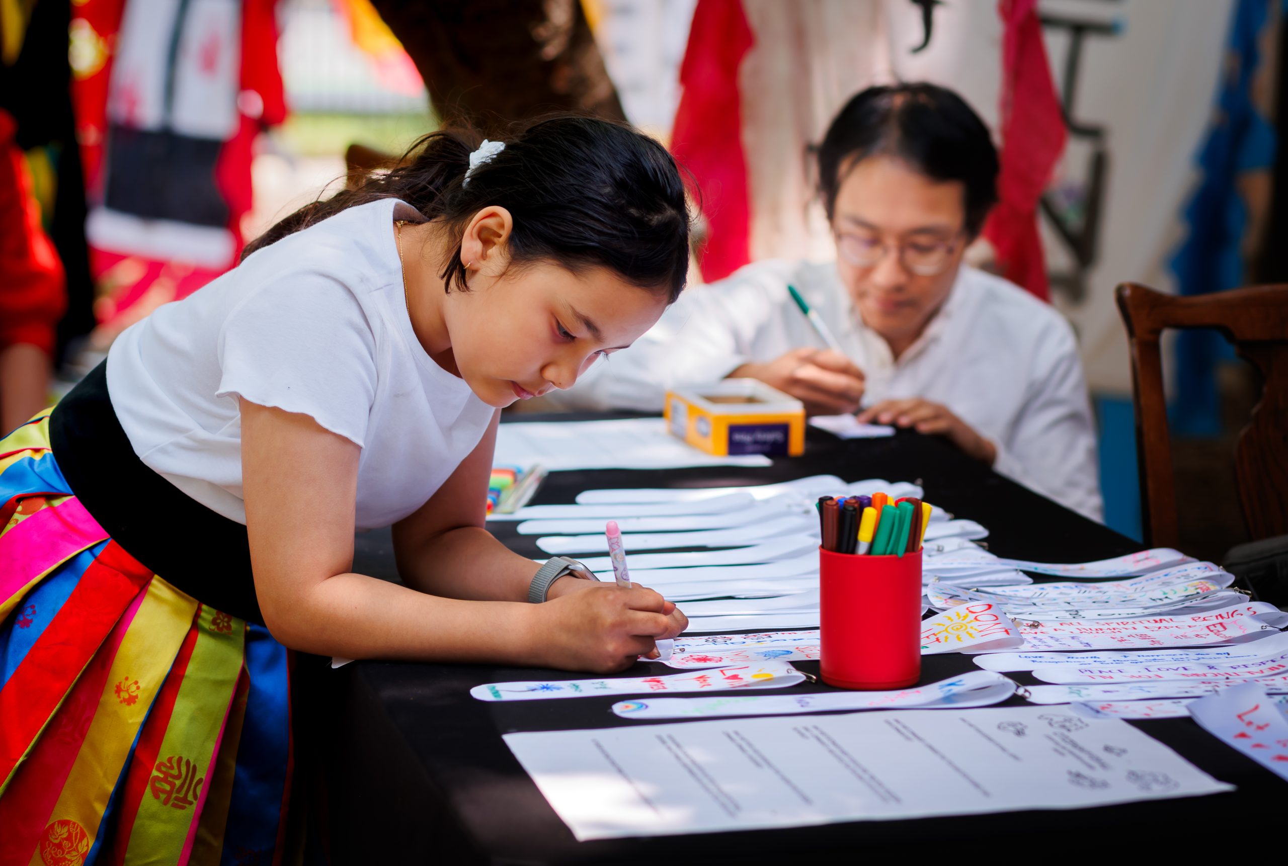 A girl in a colorful skirt leans over a table, drawing on paper with markers, while an adult in the background also writes or draws. Art supplies and decorated papers are on the table.