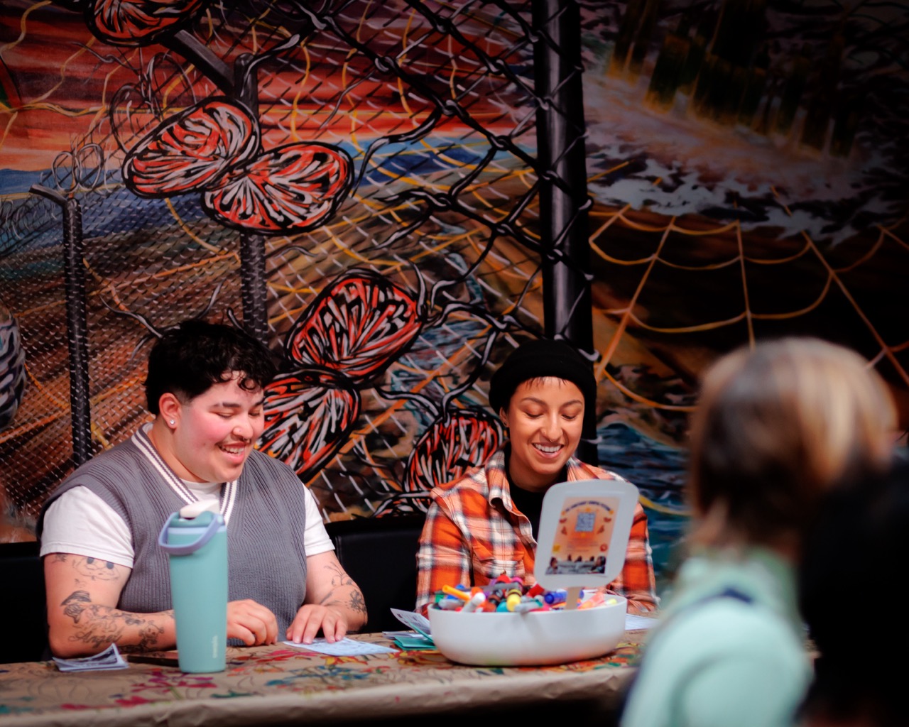 Two people sit at a decorated table, smiling and engaging with colorful materials. Behind them is a vibrant mural featuring butterflies and nature. Other figures are partially visible in the foreground.