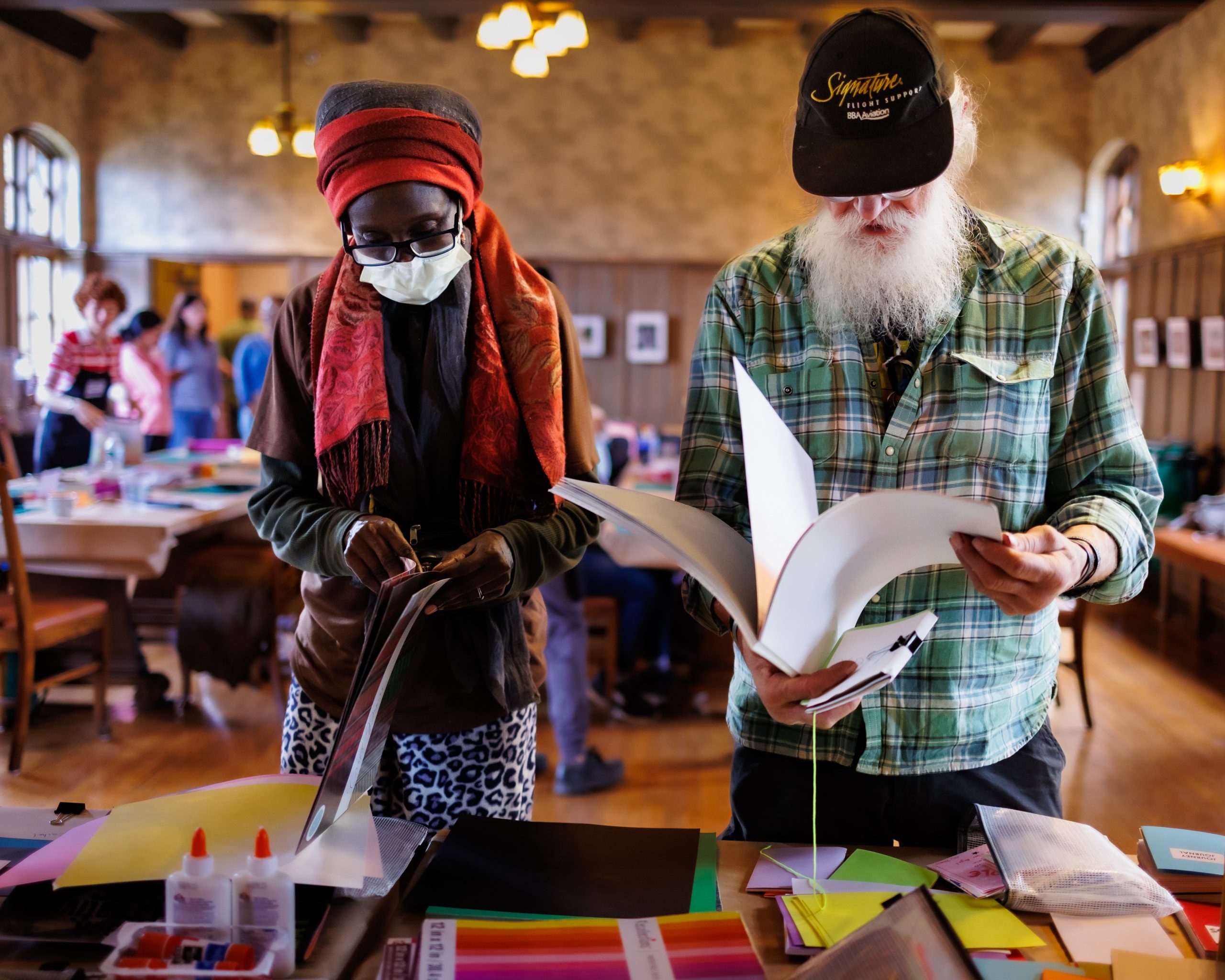 Two people stand at a table with art supplies. One, in a red scarf and face mask, handles paper, while the other, with a white beard and black cap, looks through a book. Other people work in the background of a cozy room.