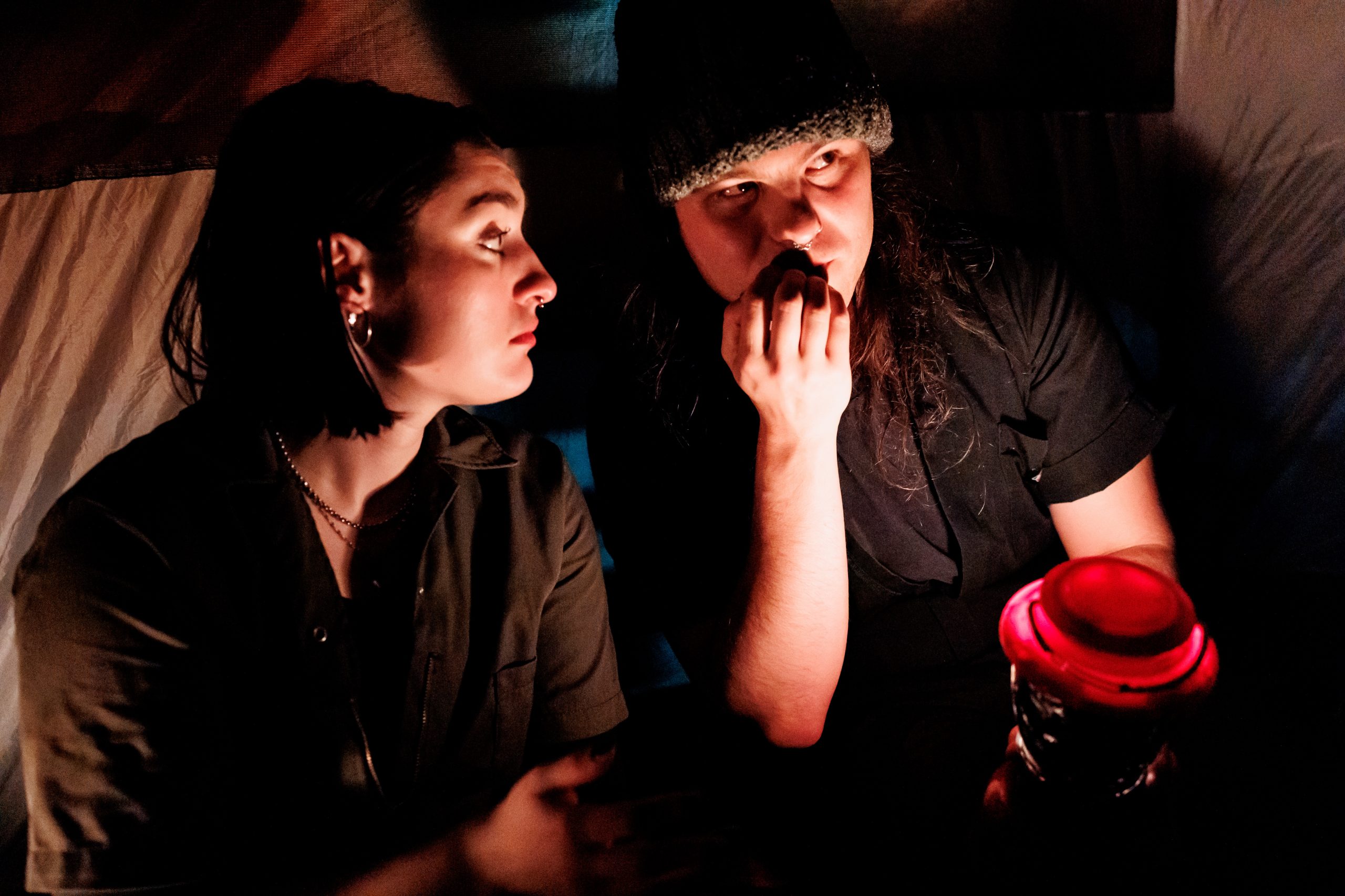 Two people sit close together in dim lighting, one holding a red lantern and looking nervous, while the other looks at them seriously, as if telling a story or comforting them.