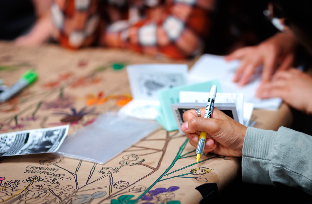 A person draws colorful flowers and leaves on a brown paper-covered table with a pen, while others nearby handle papers and booklets. The focus is on the hand holding the pen, with blurred figures in the background.