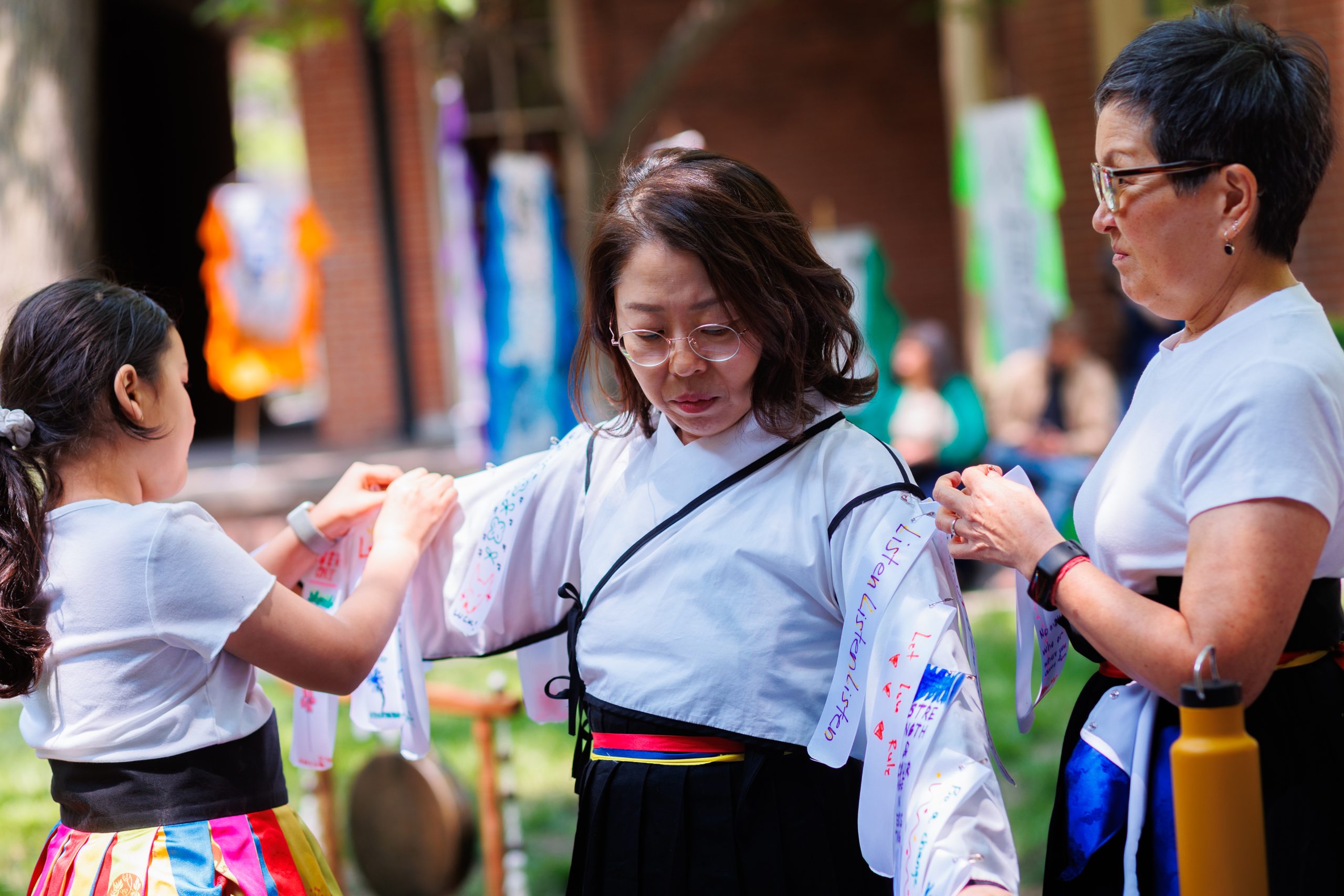 Three women outdoors help one another put on traditional clothing decorated with colorful writing, with one woman in the center adjusting her sleeves. A drum and people are visible in the blurred background.