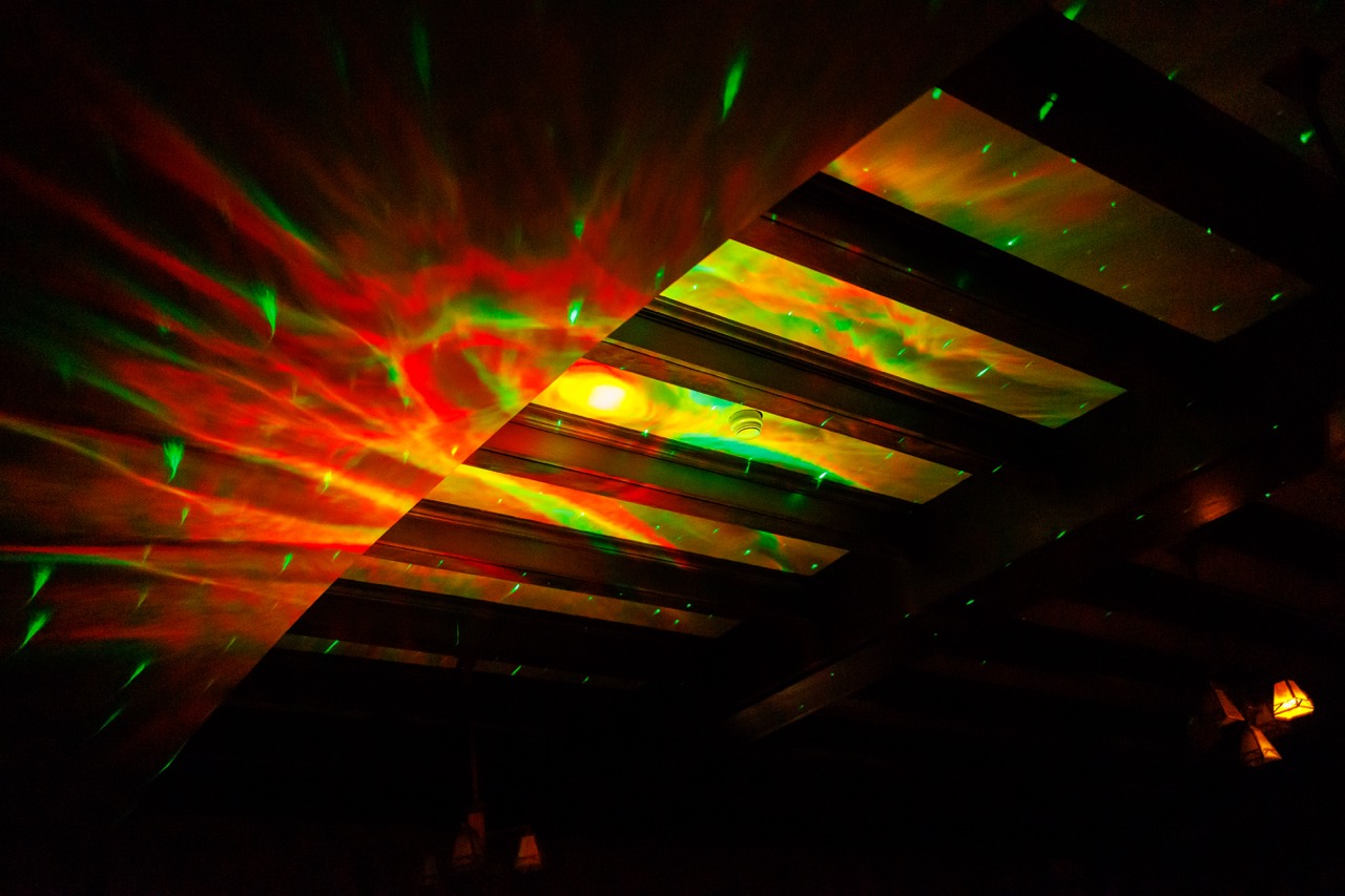 A dark ceiling with wooden beams is illuminated by vibrant red and green laser lights, creating a colorful, swirling pattern across the surface.