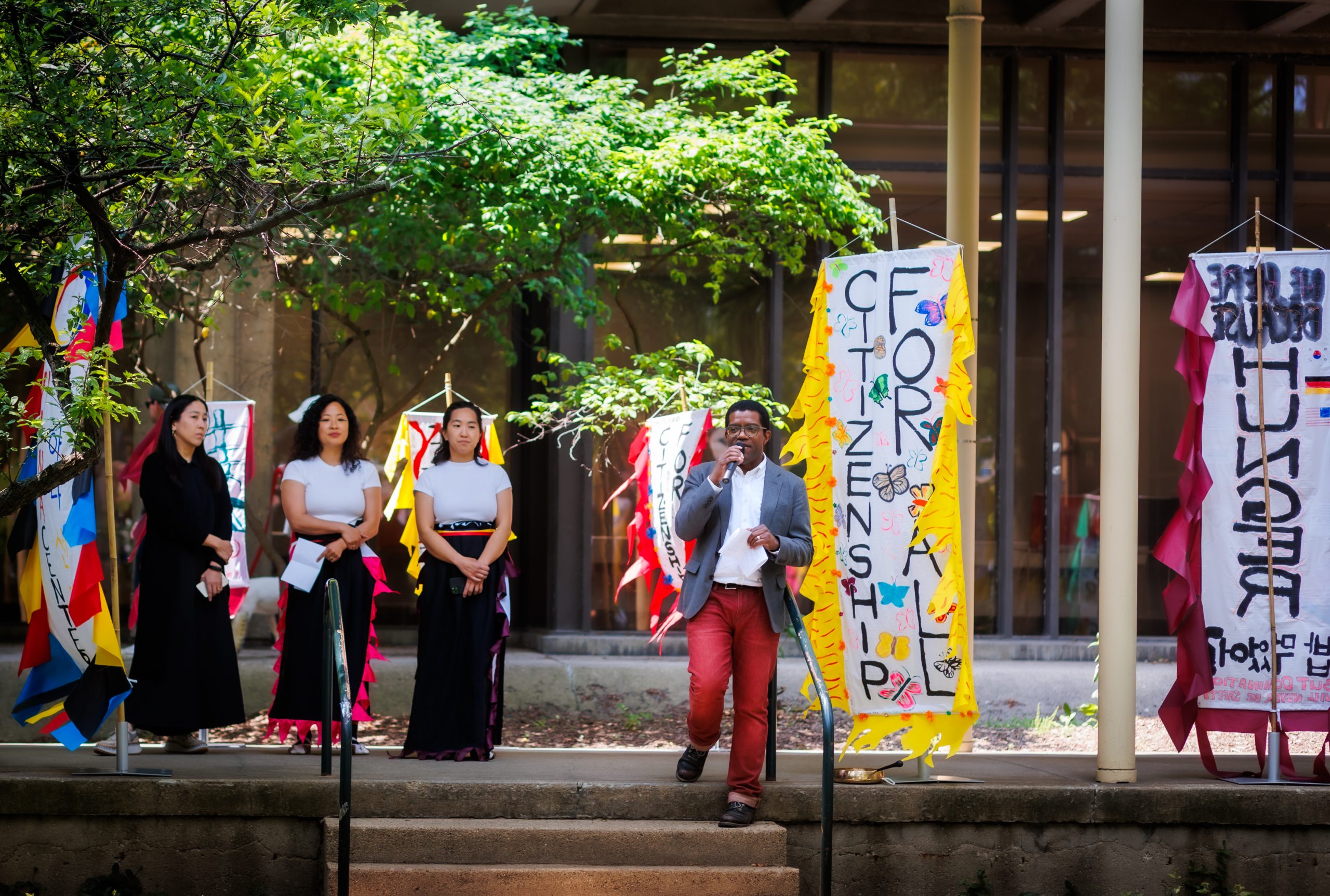 A man speaks into a microphone on outdoor steps, while three women stand behind him. Colorful banners hang nearby, and trees and a modern building are visible in the background.