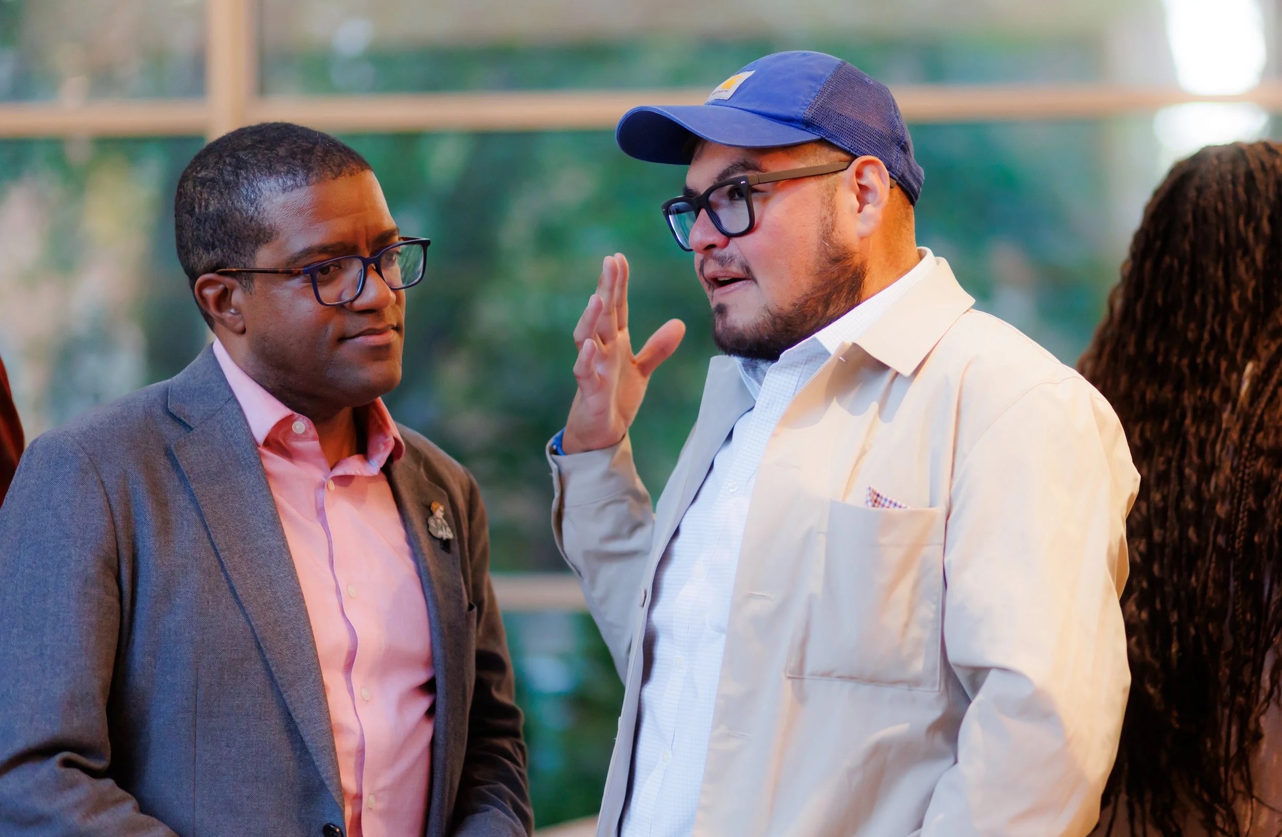 Two men are having a conversation indoors; one is wearing glasses, a grey suit, and a pink shirt, while the other wears glasses, a blue cap, and a light-colored jacket, gesturing with his hand as he speaks.