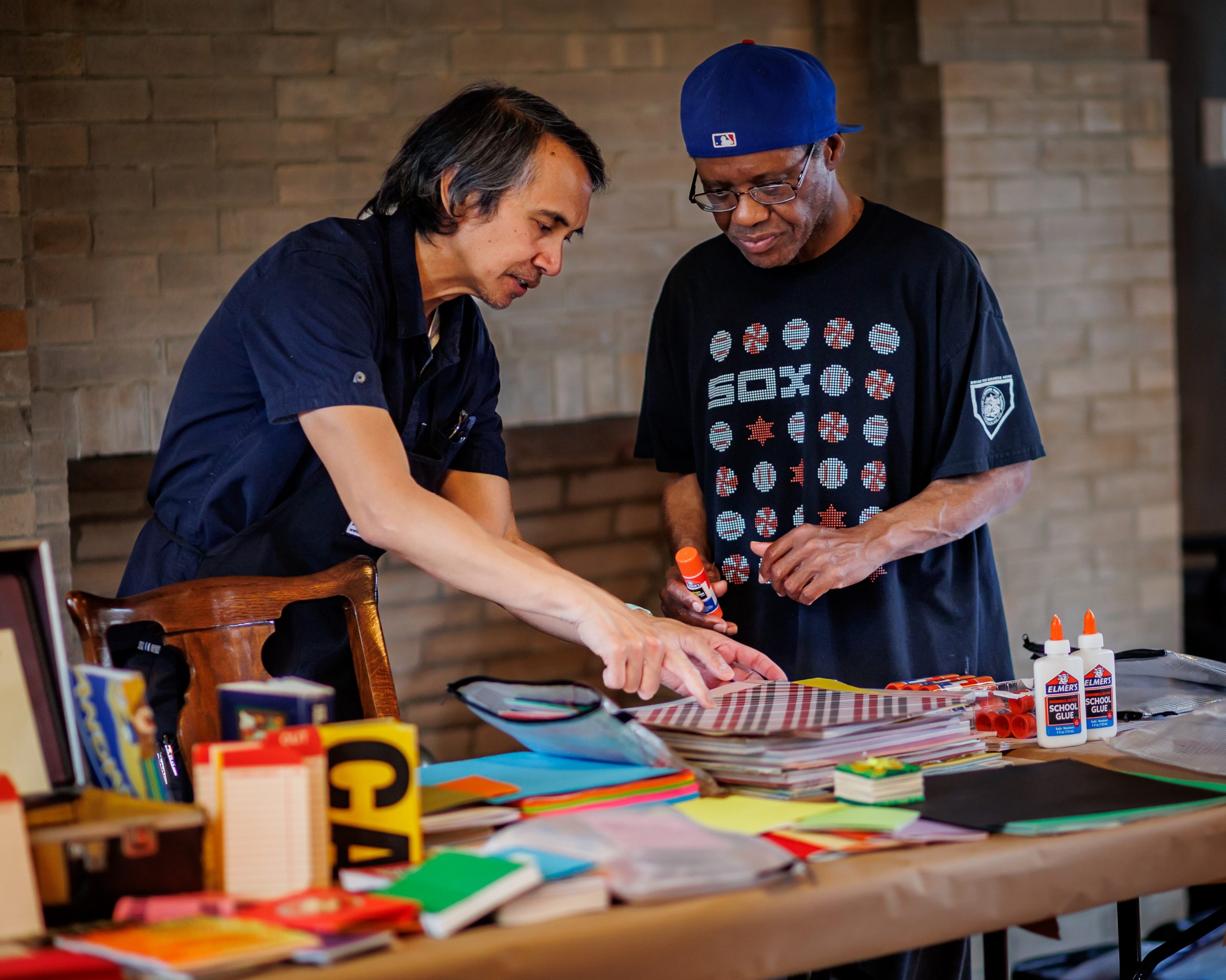 Two men stand at a table covered with craft supplies, including glue, paper, and notebooks. One man points at something on the table while the other holds a glue stick, both focusing on a creative project.