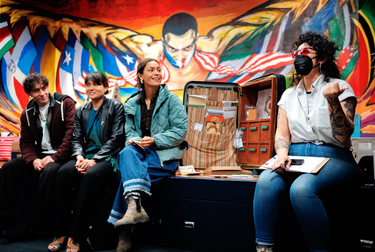 Four people sit and chat in front of a colorful mural with flags and a powerful figure. Three are seated together, smiling and listening to a fourth person, who wears a mask and holds a clipboard next to an open suitcase.
