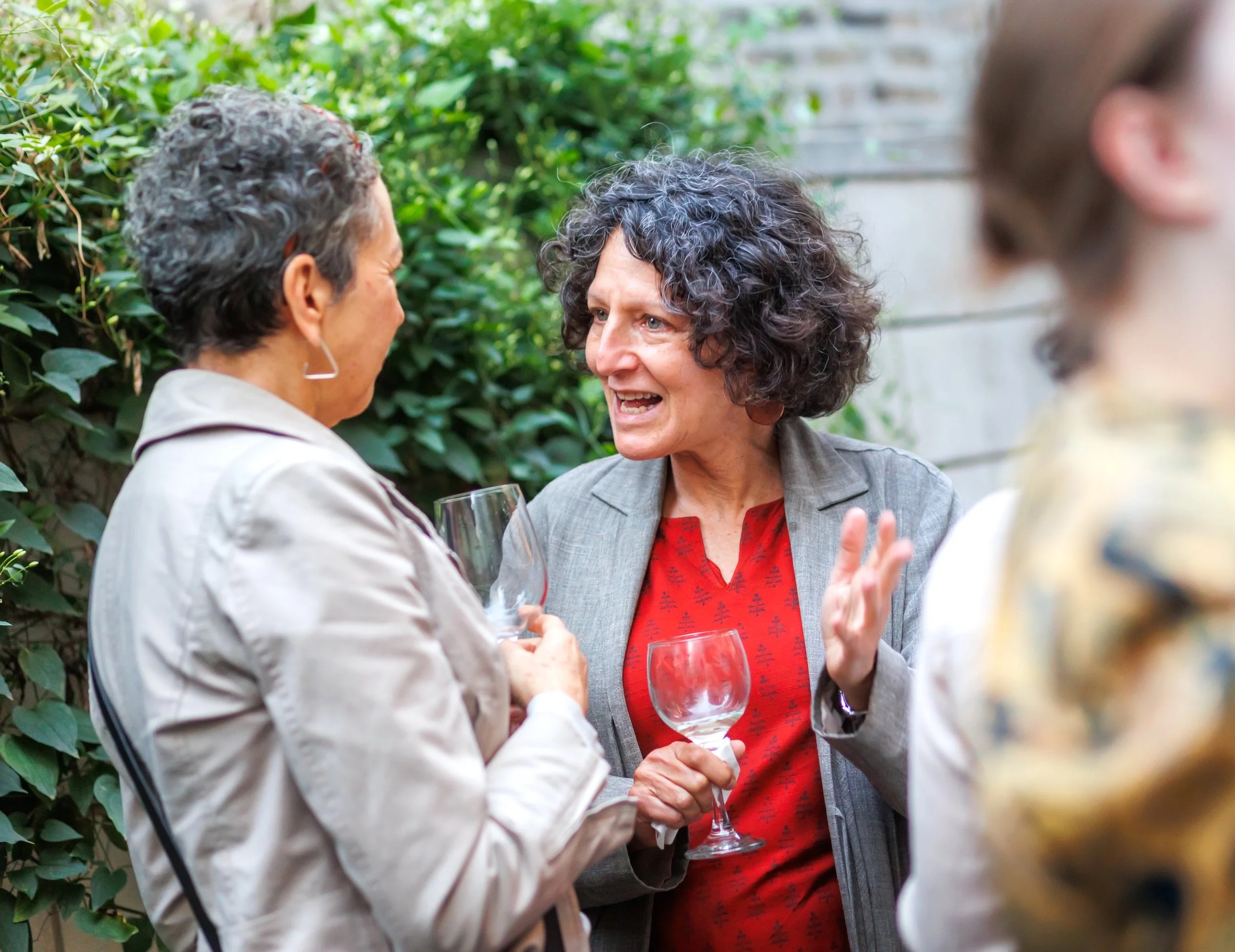 Two women stand outdoors, smiling and conversing while holding empty wine glasses. Greenery and a brick wall are in the background, and another person is partially visible in the foreground.