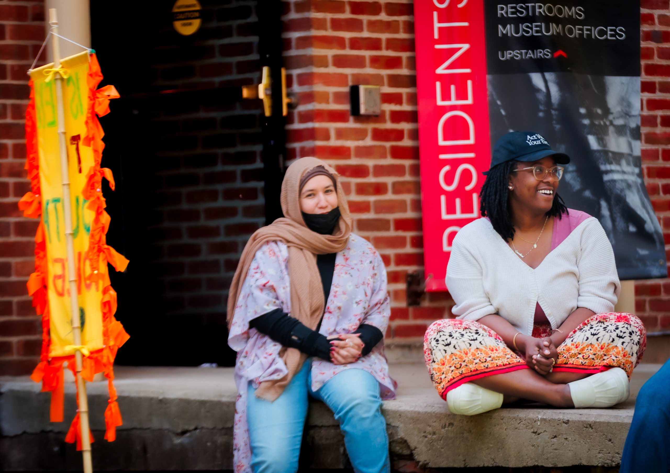 Two women sit on a low brick wall. One wears a hijab and holds a black mask under her chin; the other wears glasses and a ball cap. A bright yellow sign and restroom sign are visible in the background.