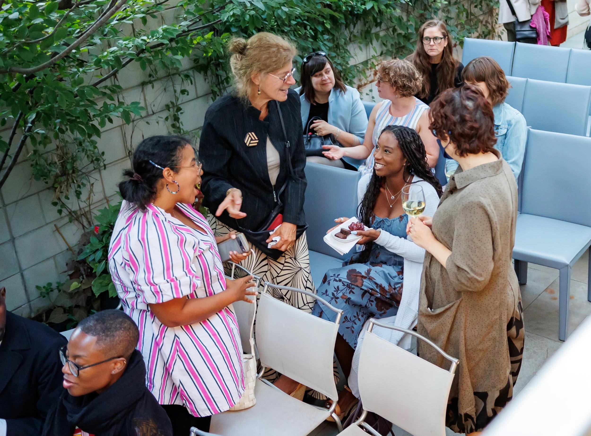 A group of women, some seated and some standing, chat and smile together at an outdoor event with rows of light blue chairs and greenery in the background. One woman holds a glass of wine.