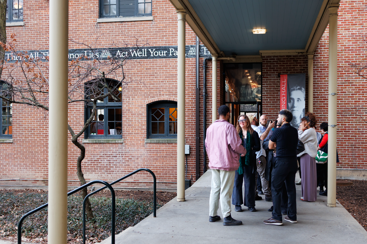 A group of people stand and talk outside a brick building under a covered walkway. Above the entrance, a quote reads, There All the Honor Lies—Act Well Your Part. A large vertical banner with a face is mounted nearby.
