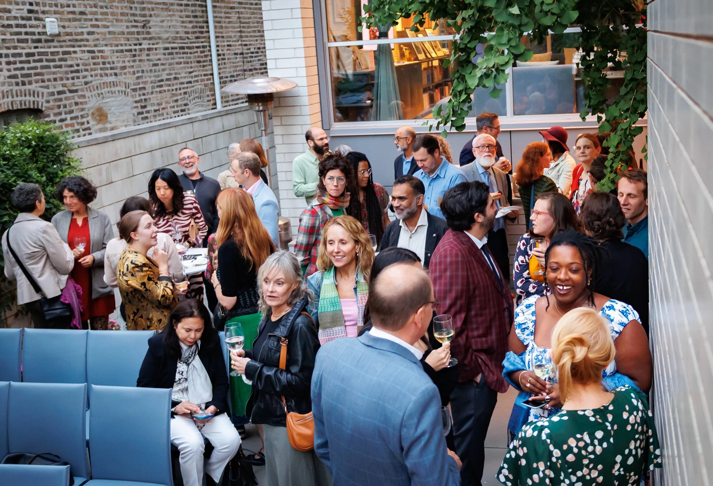 A diverse group of people mingle and converse at an outdoor social event, holding drinks and standing close together, surrounded by brick walls and greenery.