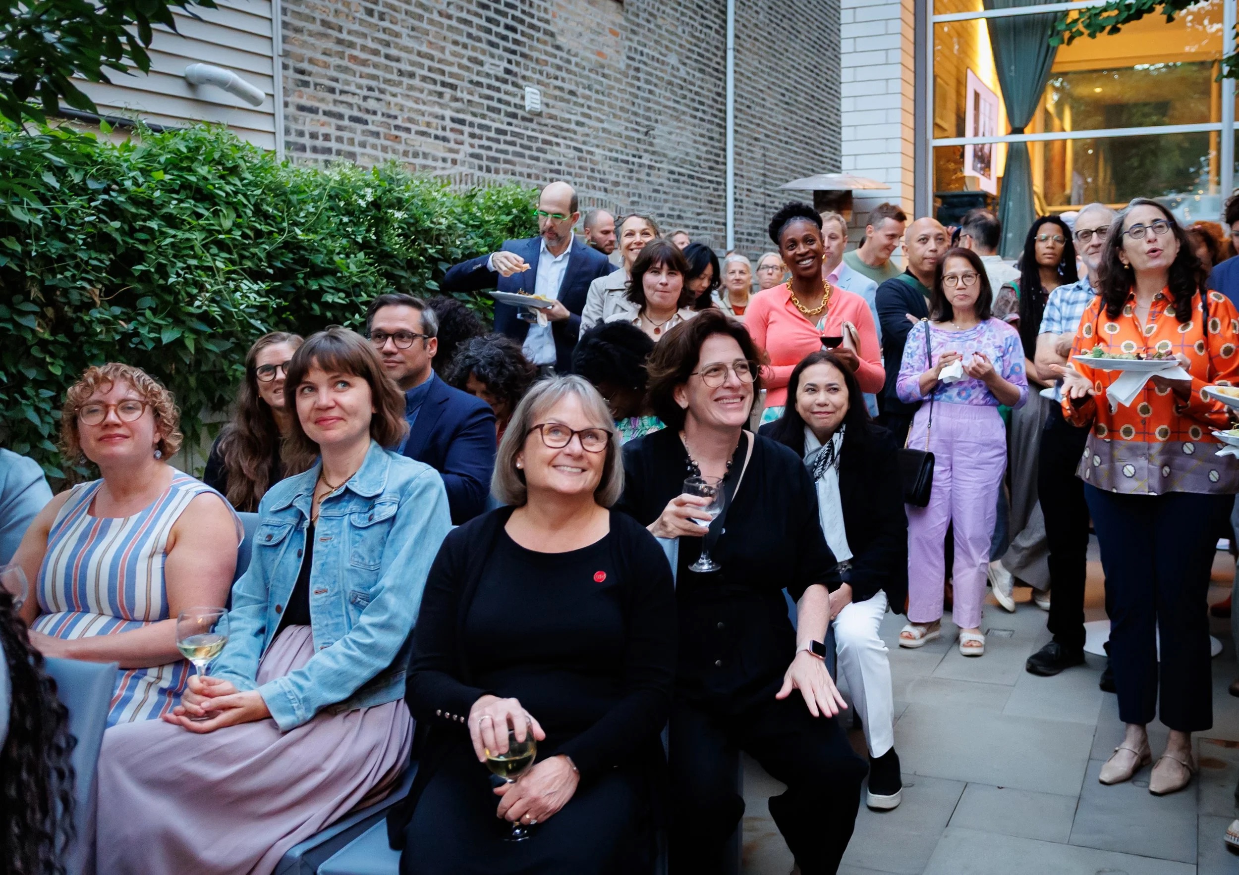 A diverse group of people, some seated and some standing, gather outdoors at an event. Many are smiling, holding drinks, and facing the camera. Greenery and brick walls surround the patio area.