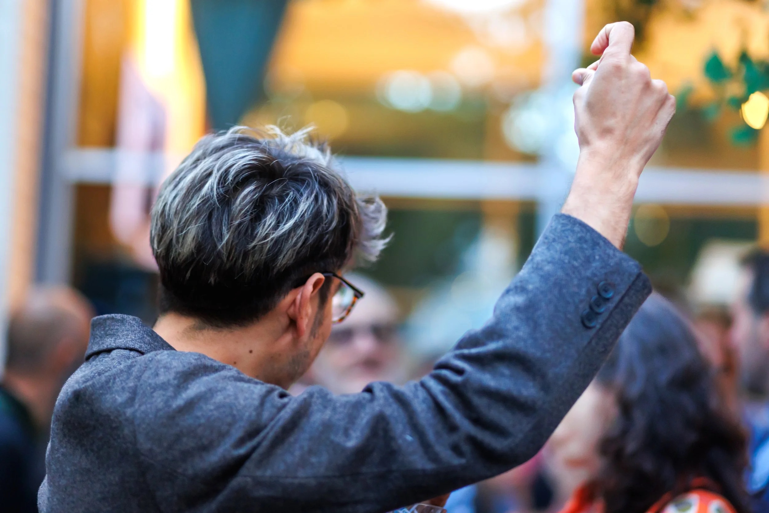 A person with short, dark hair with gray highlights, wearing glasses and a gray jacket, raises their right arm in the air. The background is blurred with several people and outdoor elements.