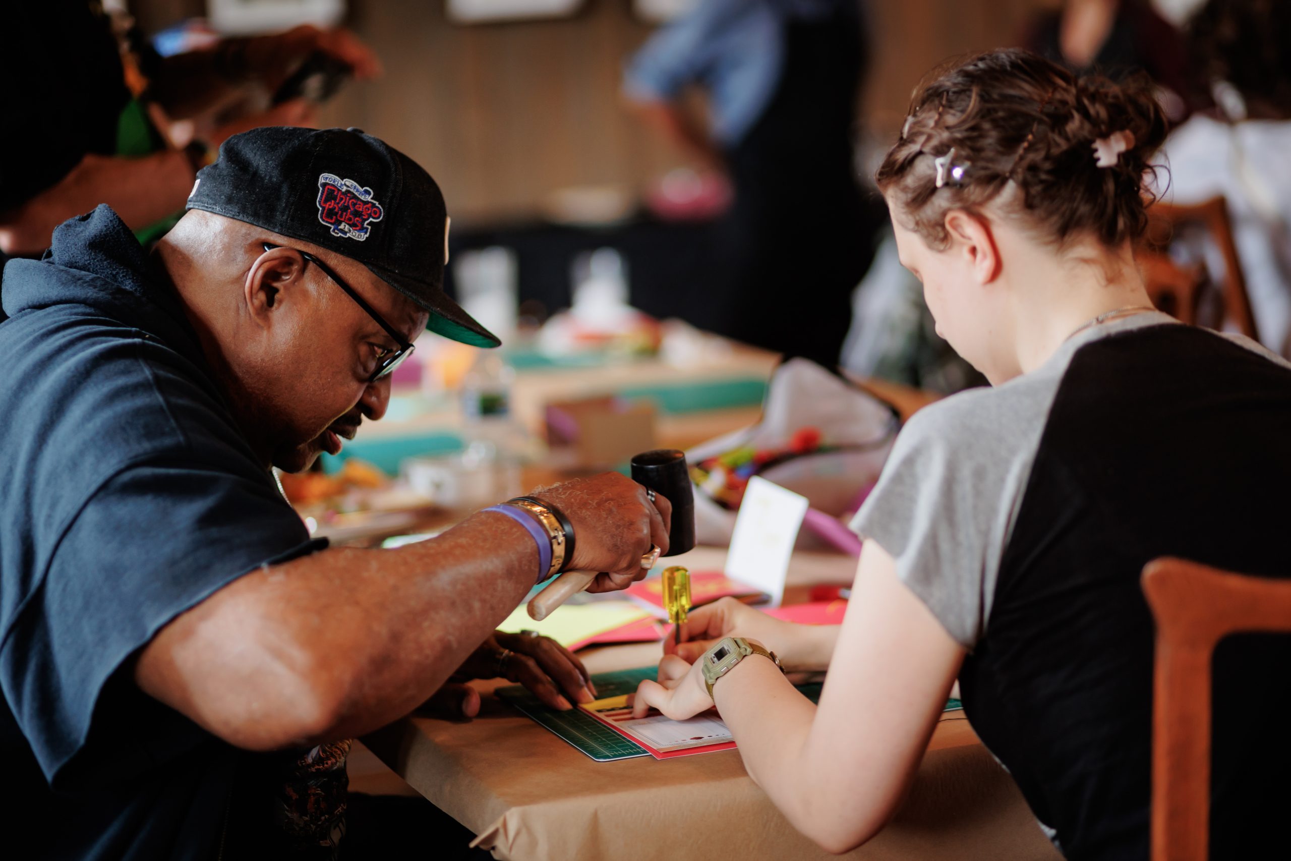 A man and a woman sit at a table covered with colorful craft supplies, focused on creating art together. The man uses a tool, while the woman draws or writes, surrounded by a busy, creative environment.
