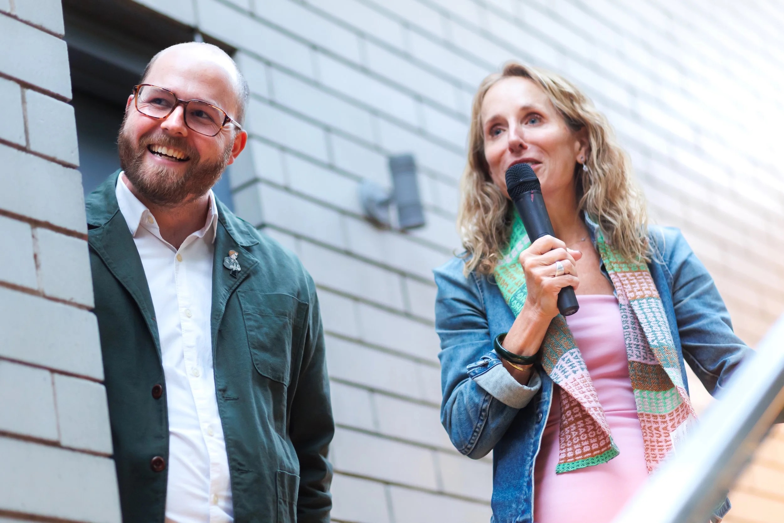 A smiling man in glasses and a green jacket stands next to a woman holding a microphone, speaking. She wears a pink dress, a denim jacket, and a colorful scarf. They are outdoors in front of a light brick wall.