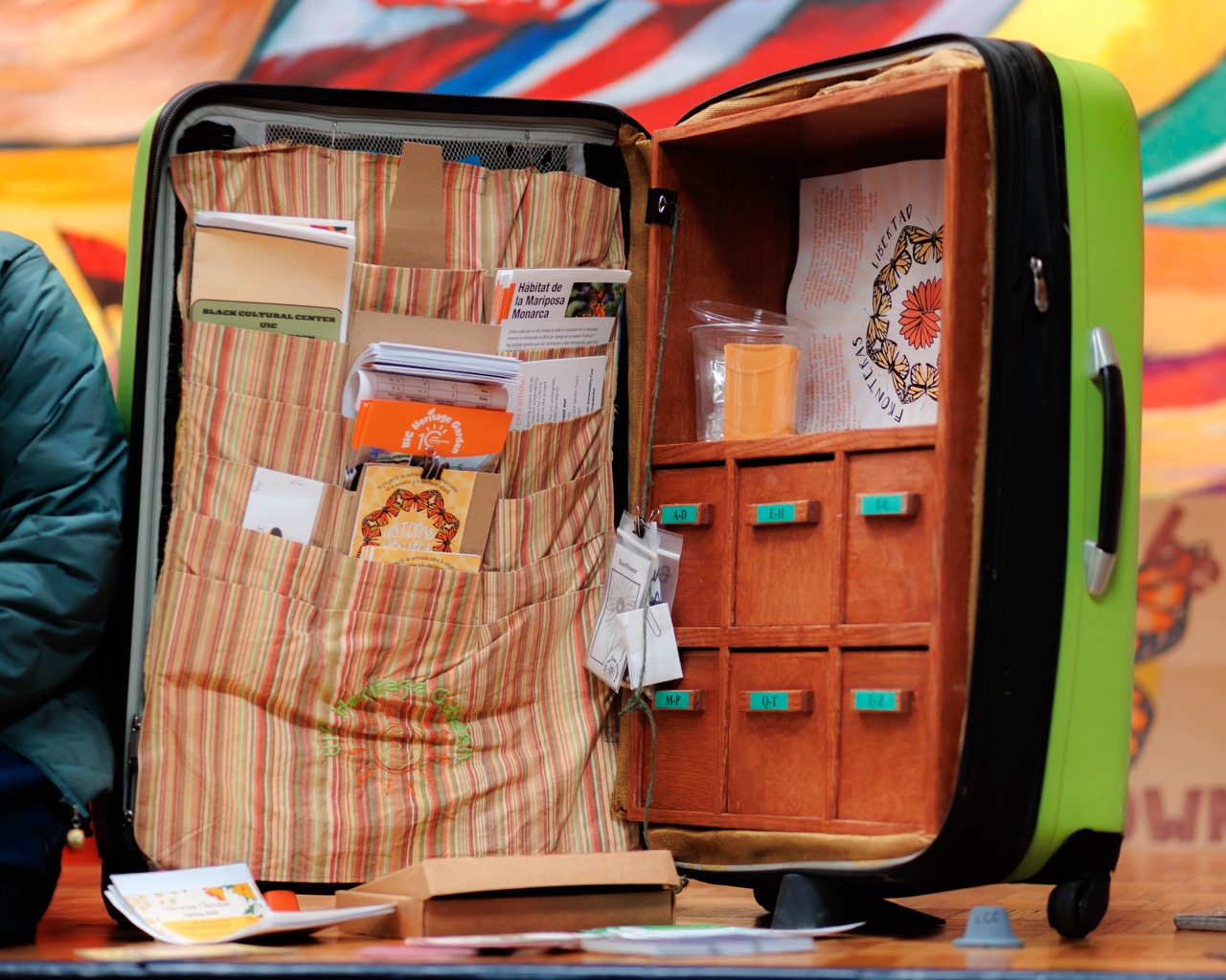 A green suitcase stands open on a table, with one side holding various booklets, brochures, and papers in striped pockets, and the other side containing small drawers and compartments, some labeled and one holding a clear plastic cup.
