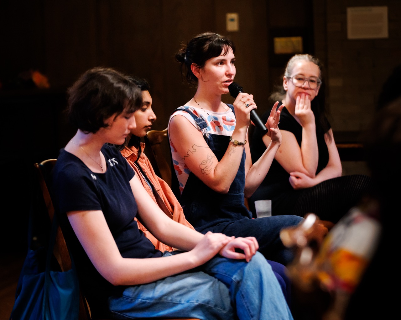 Four people sit in a row, with one person in the center holding a microphone and speaking. The others listen attentively. The setting appears to be an indoor event or discussion panel.
