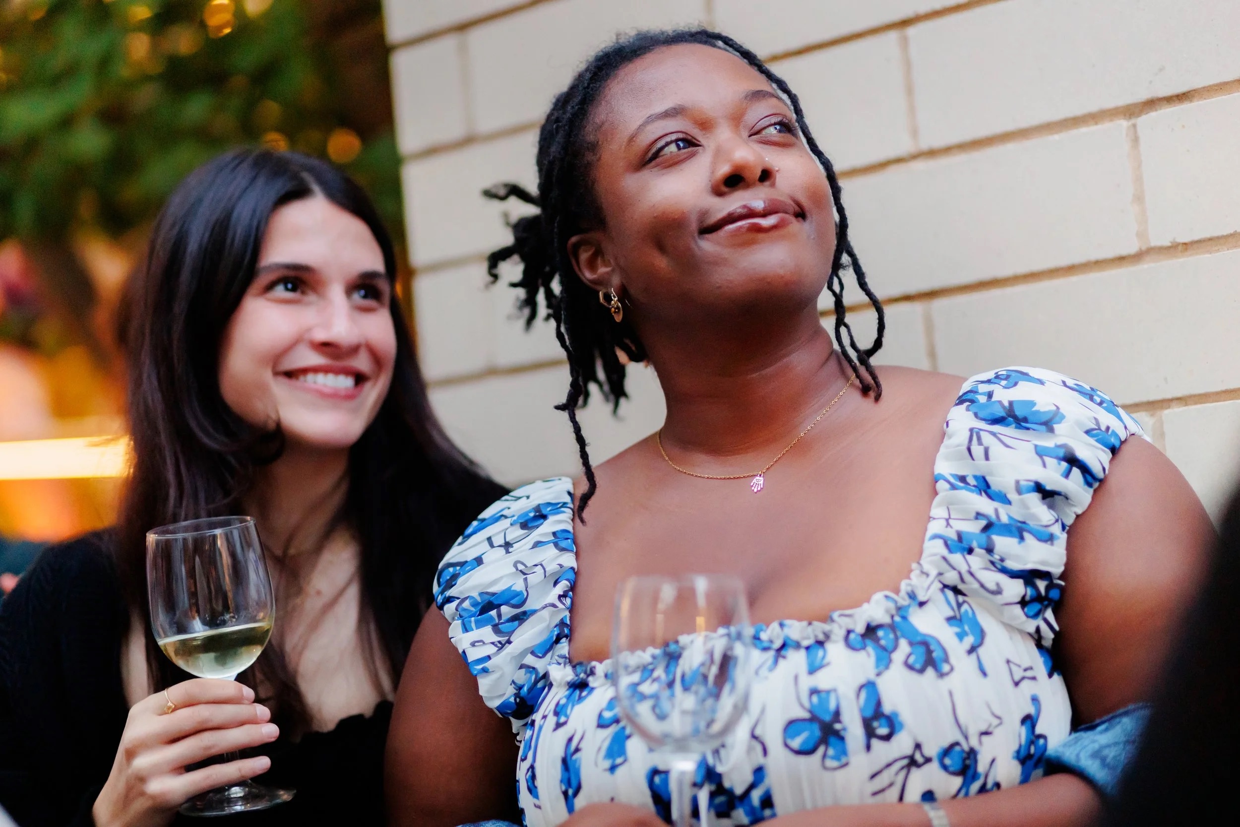 Two women smiling and holding glasses of white wine at an outdoor gathering. One wears a white dress with a blue butterfly pattern, the other has long dark hair and wears black. They appear happy and relaxed.