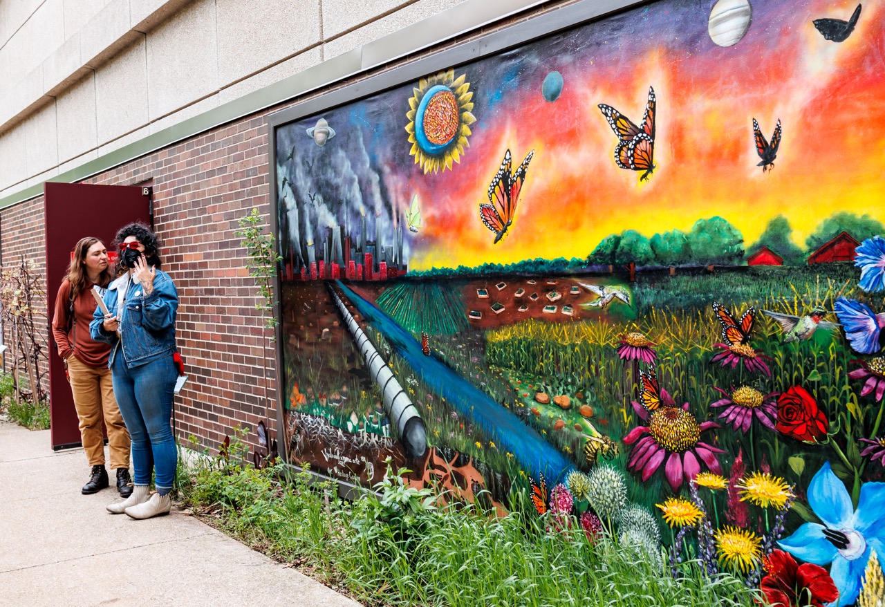 Two people stand next to a vibrant mural on a brick wall, depicting a colorful landscape with flowers, butterflies, pollution, and planets, contrasting nature with industrial elements.