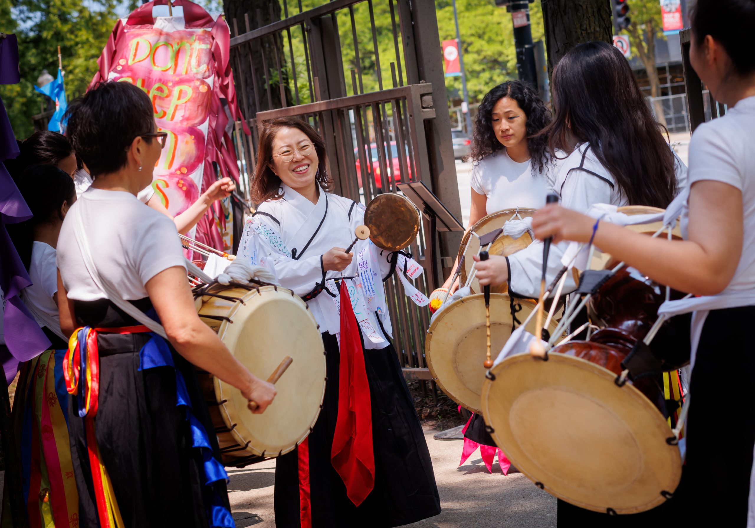 A group of women wearing traditional Korean clothing play drums together outdoors, smiling and enjoying the activity near a metal fence and colorful decorations.