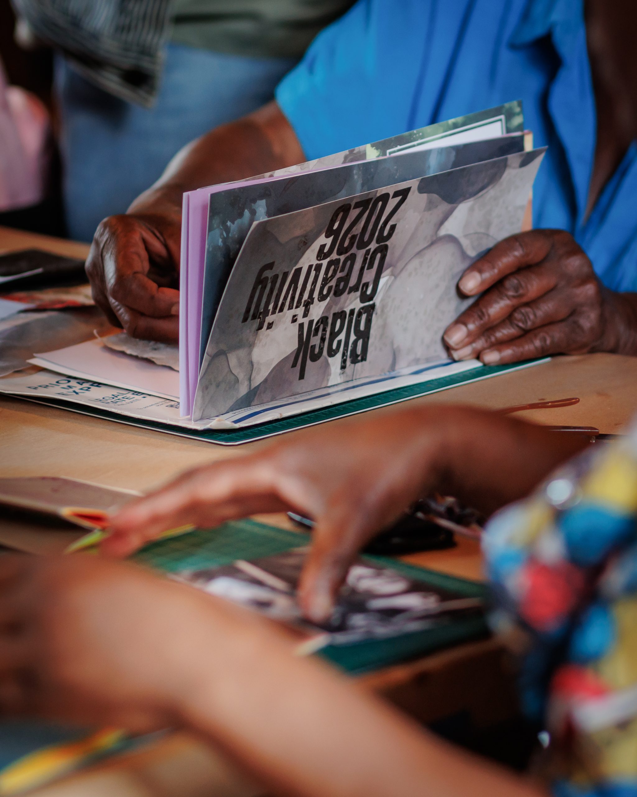 Two people sit at a table working with paper and photos. One person holds an upside-down booklet that reads Black Creativity 2022. Their hands and colorful clothing are visible in the foreground.