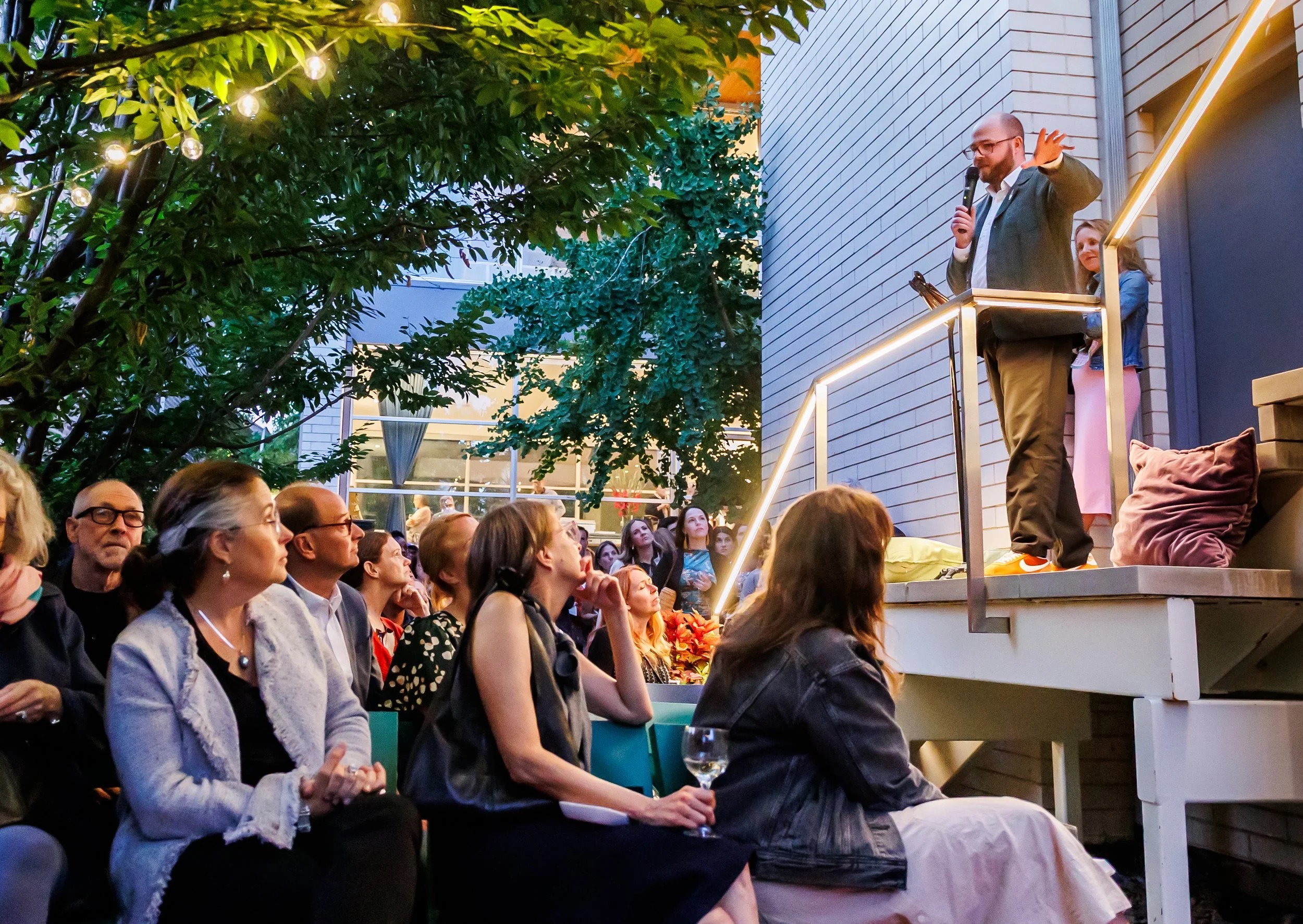 A man speaks into a microphone on an outdoor patio steps at night, addressing a seated crowd under string lights, with trees and a modern building in the background.