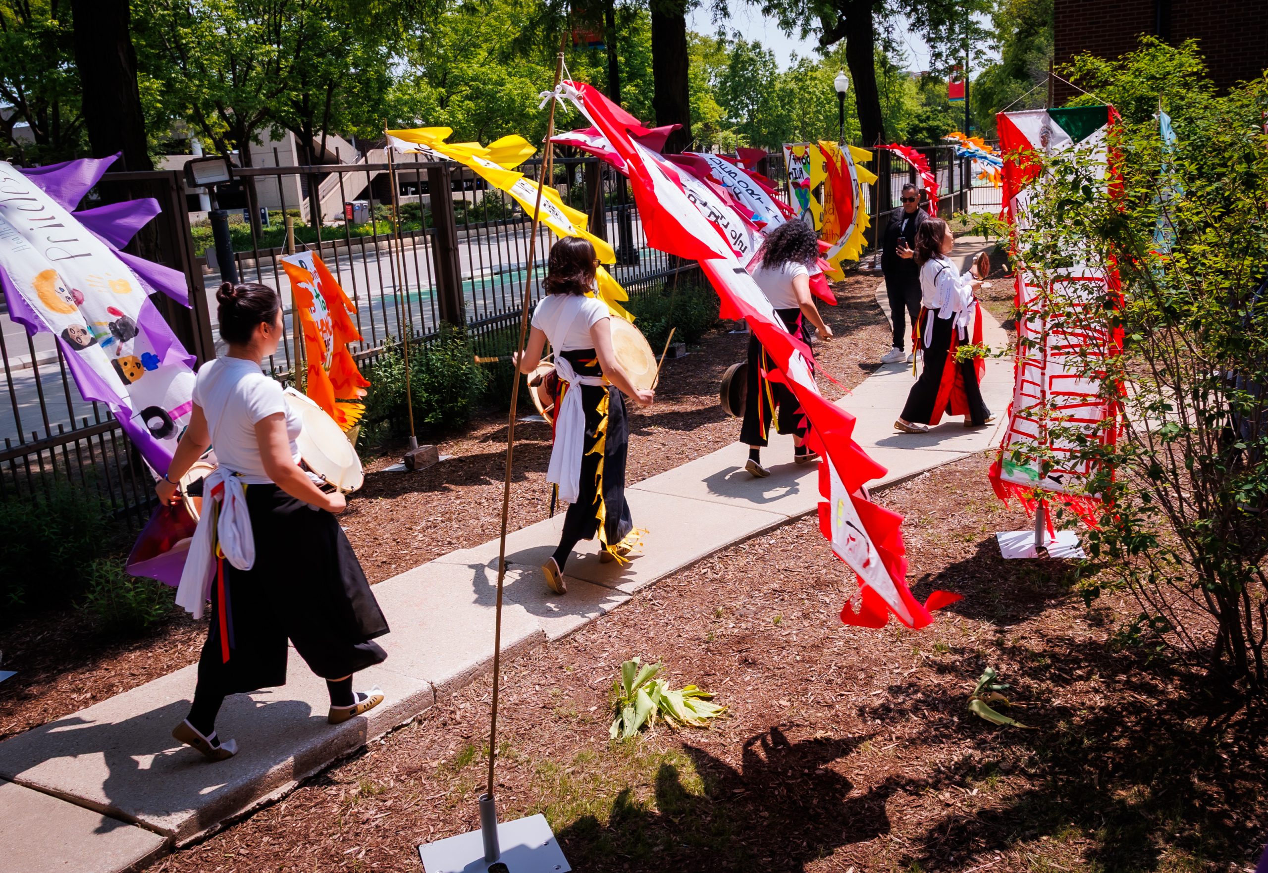 People wearing traditional clothing walk along a sidewalk decorated with colorful flags and banners, with trees and a metal fence in the background on a sunny day.