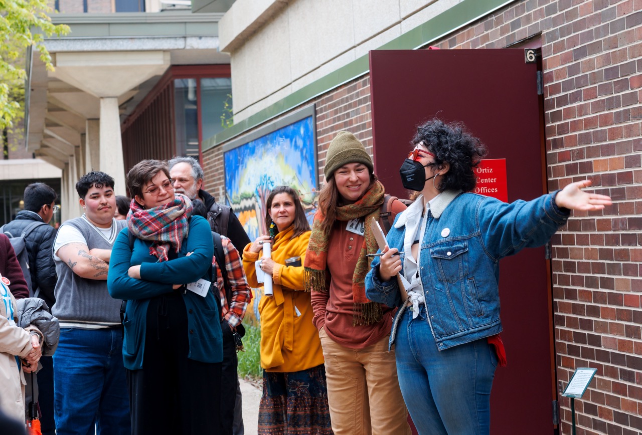 A group of people stands outside a brick building, some smiling and listening as a person in a denim jacket and face mask gestures while speaking. Several people hold papers, and colorful artwork is displayed on the wall behind them.