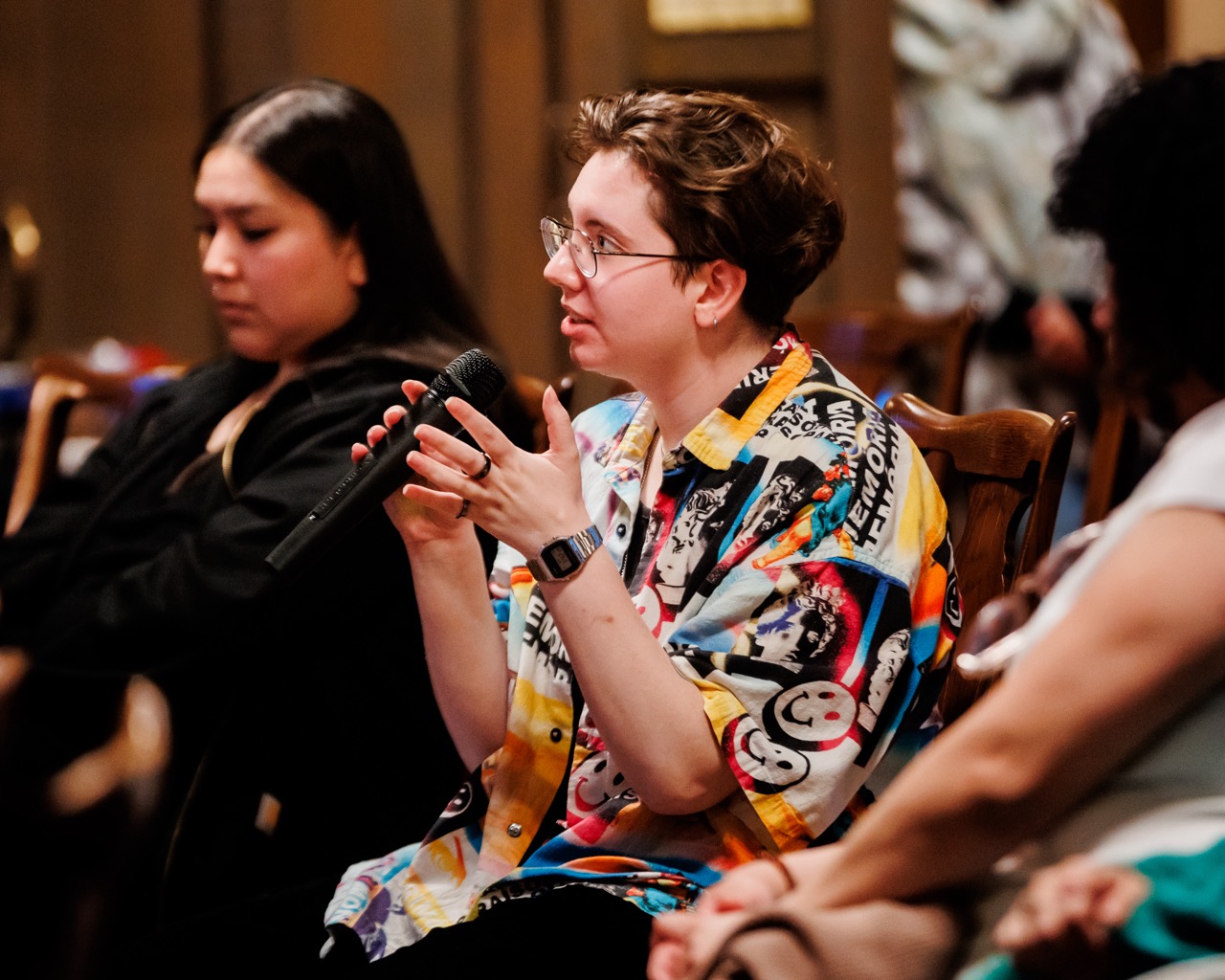 A person wearing glasses and a colorful shirt holds a microphone while speaking, seated among others in an indoor setting. Two other people are visible, listening attentively.