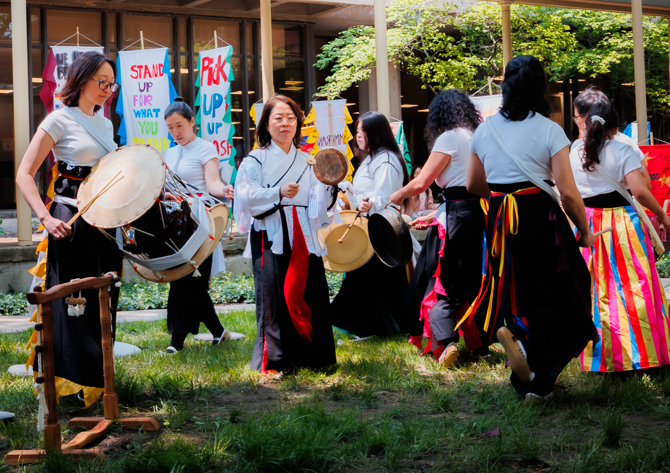 A group of women in traditional attire play drums and dance outdoors near colorful banners that read “STAND UP FOR WHAT YOU BELIEVE IN” and “RISE UP.” Sunlight filters through trees in the background.
