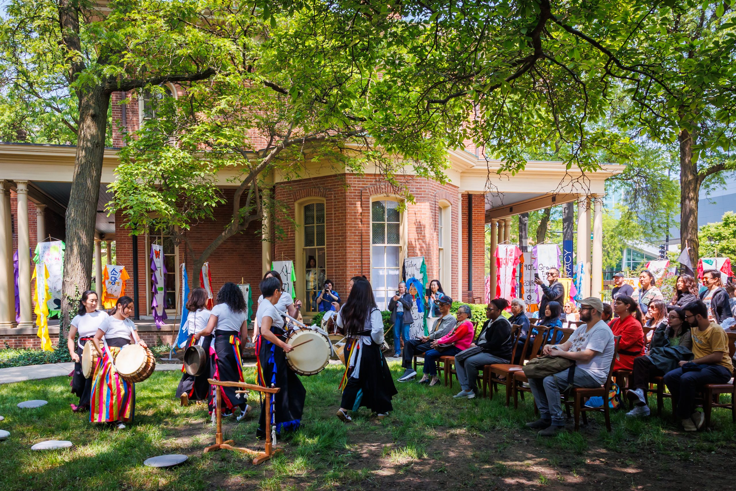 A group of performers in colorful skirts play drums outside a brick building as an audience watches. Bright banners hang from the porch, and trees provide shade over the lively outdoor event.