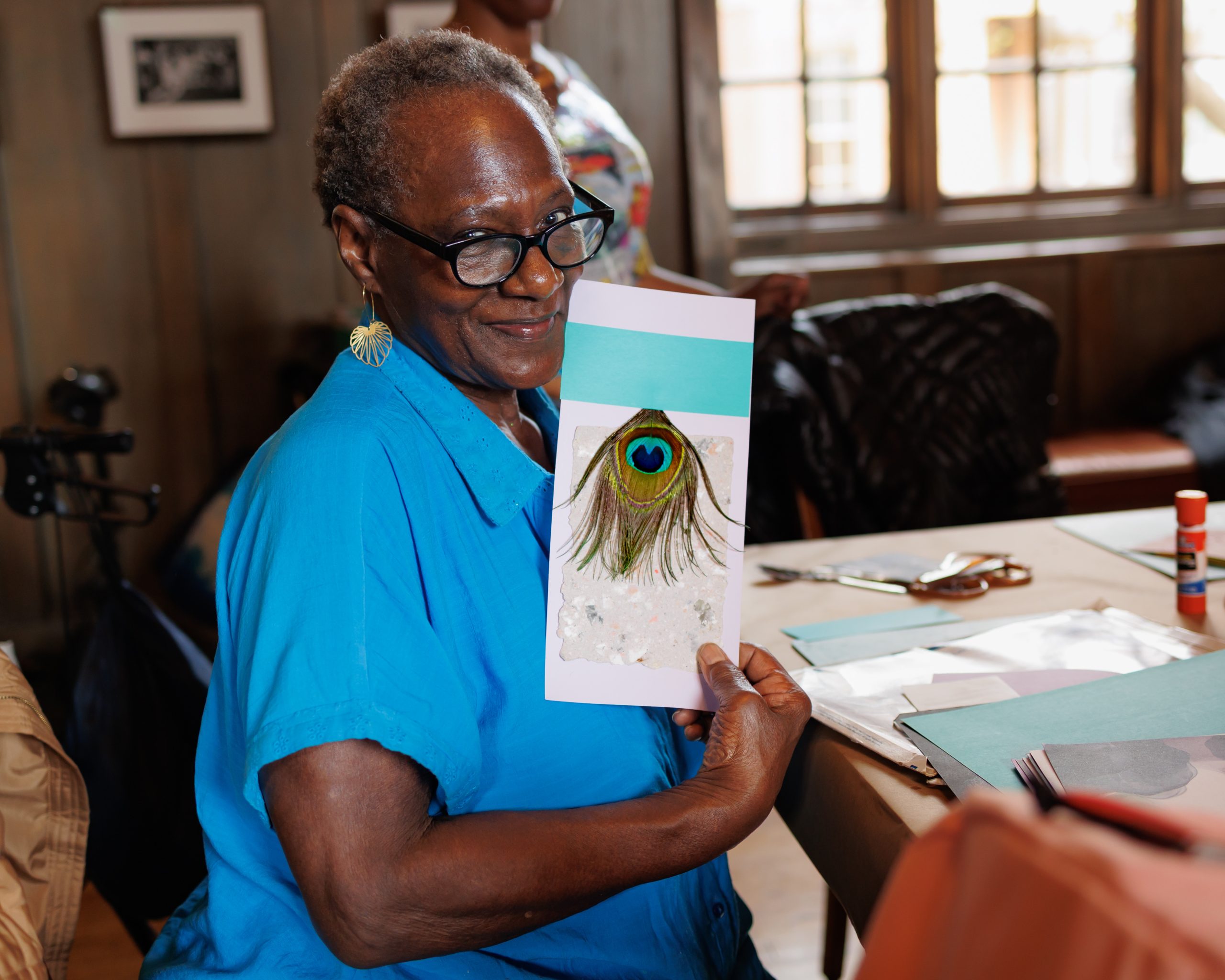 An older woman wearing glasses and a blue shirt smiles while holding up a handmade card decorated with a peacock feather. She is seated at a table with art supplies in a well-lit room.
