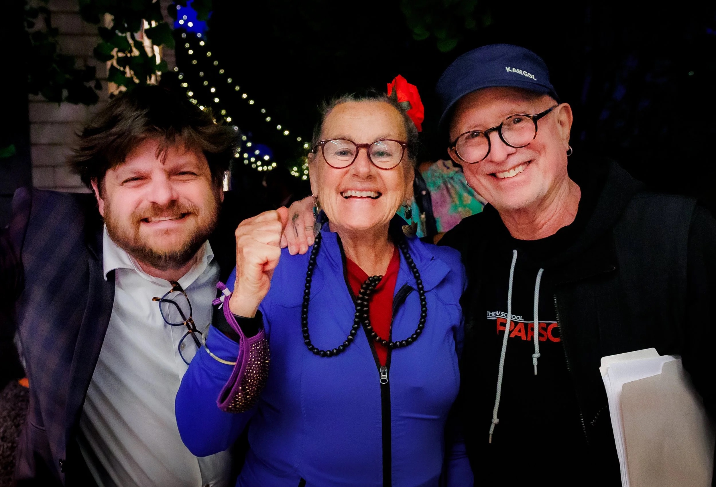 Three people are smiling and posing together outdoors at night. The person in the middle is wearing glasses, a purple jacket, and a red flower in their hair. String lights are visible in the background.