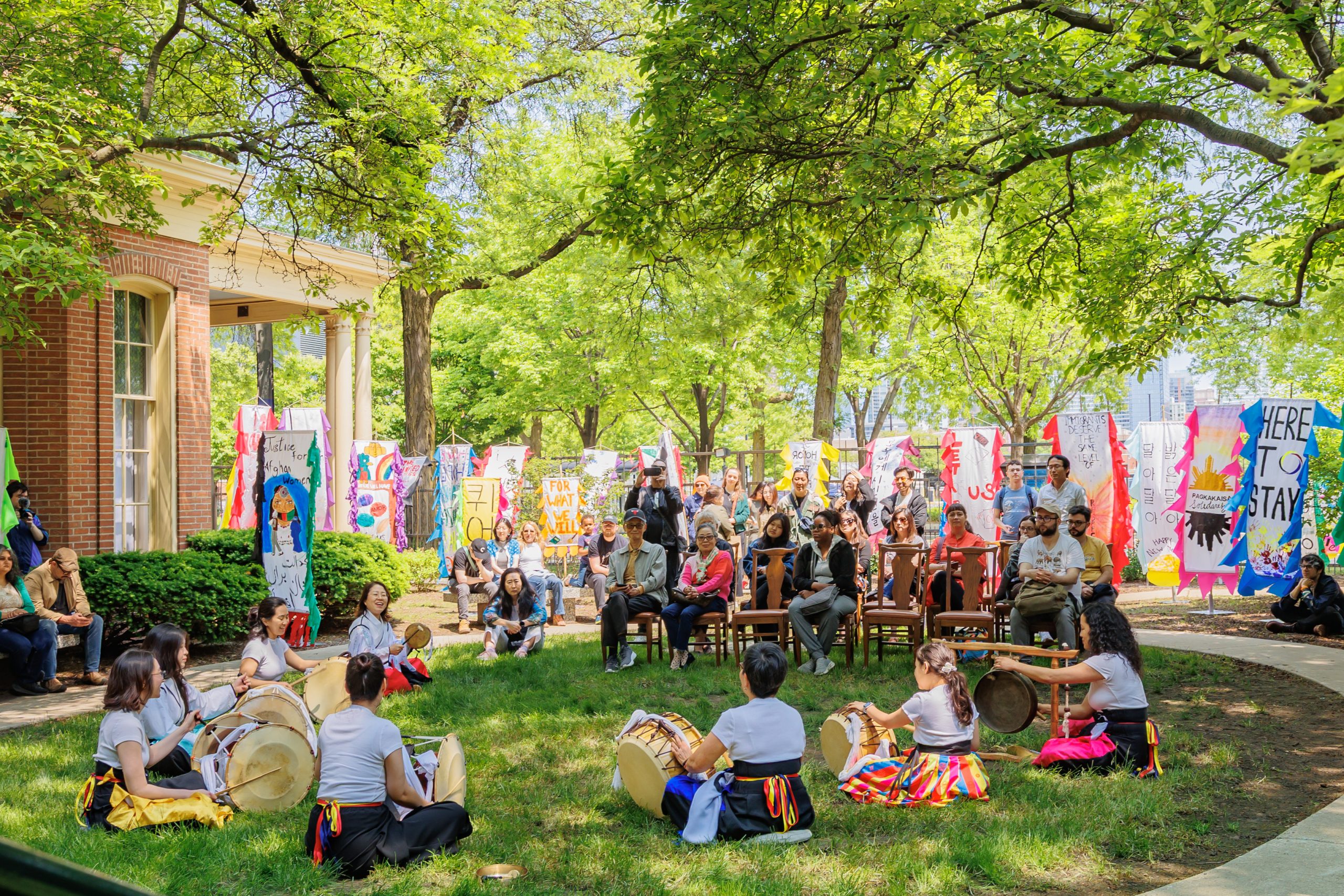 A group of people watches musicians playing drums in a sunny outdoor area surrounded by colorful banners, trees, and greenery. Some people are seated while others stand among the art displays.