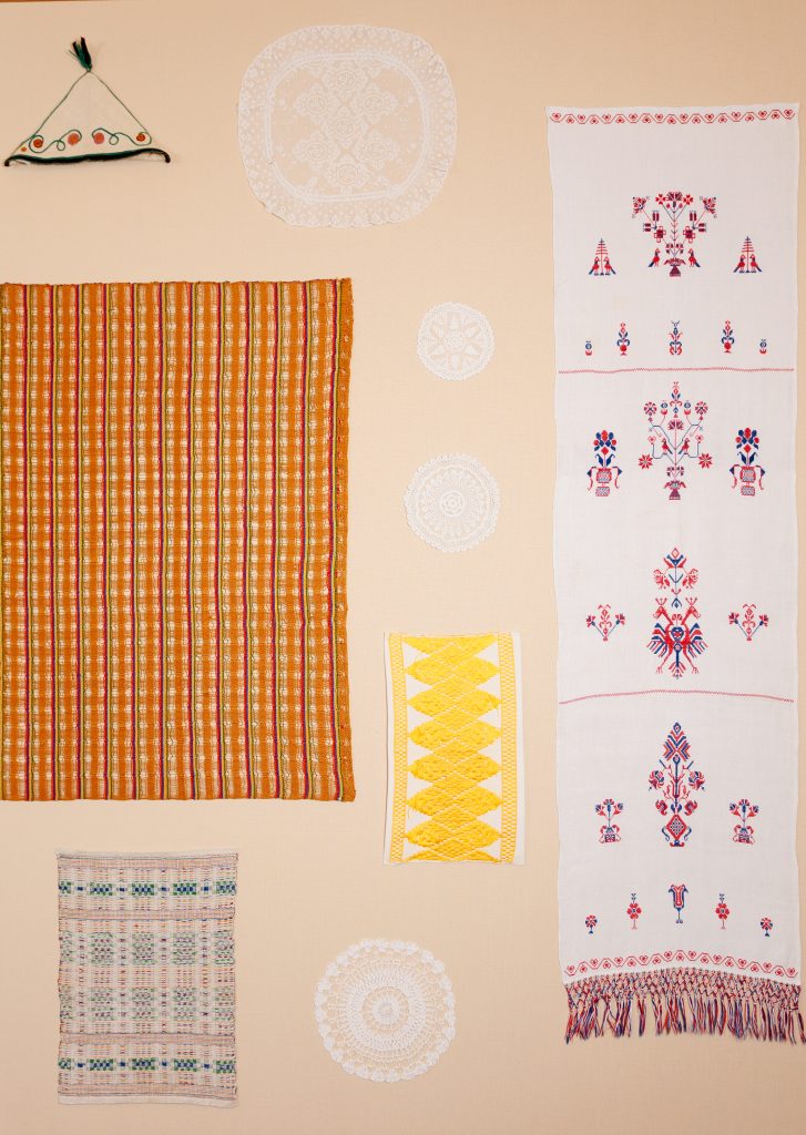 A display of colorful patterned textiles, including striped, embroidered, and crocheted cloths, arranged on a beige wall with circular lace doilies and a small triangular green decorative piece.