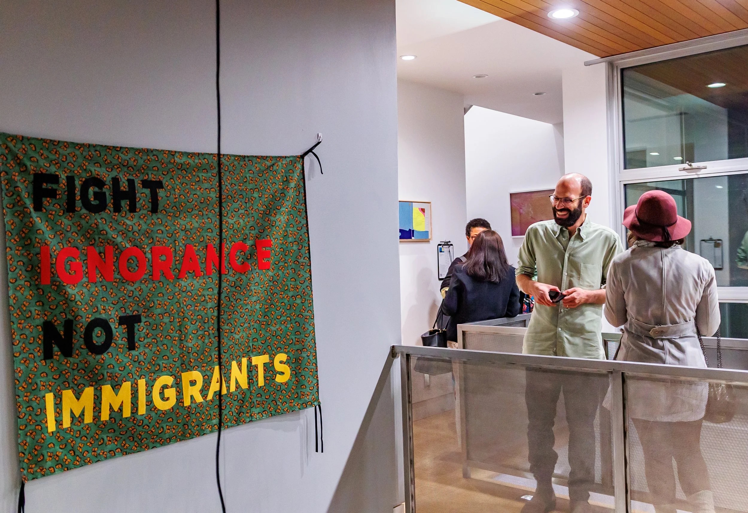 A colorful sign on a wall reads FIGHT IGNORANCE NOT IMMIGRANTS. In the background, people are talking in a modern indoor space with artwork on the walls.