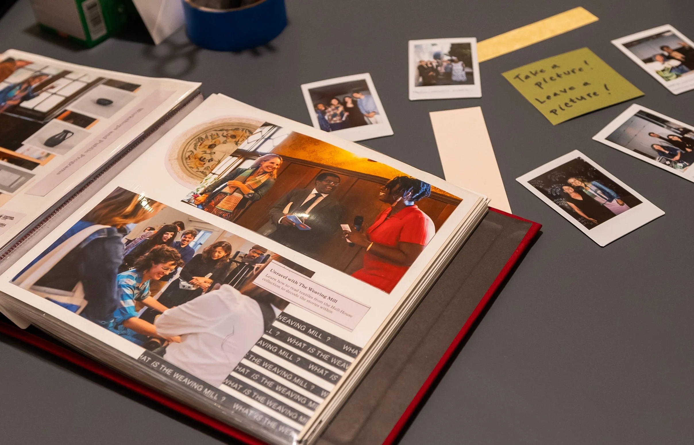 An open photo album with candid event photos sits on a table, surrounded by instant photos and a note reading, “Take a picture, leave a picture!” on a yellow card.