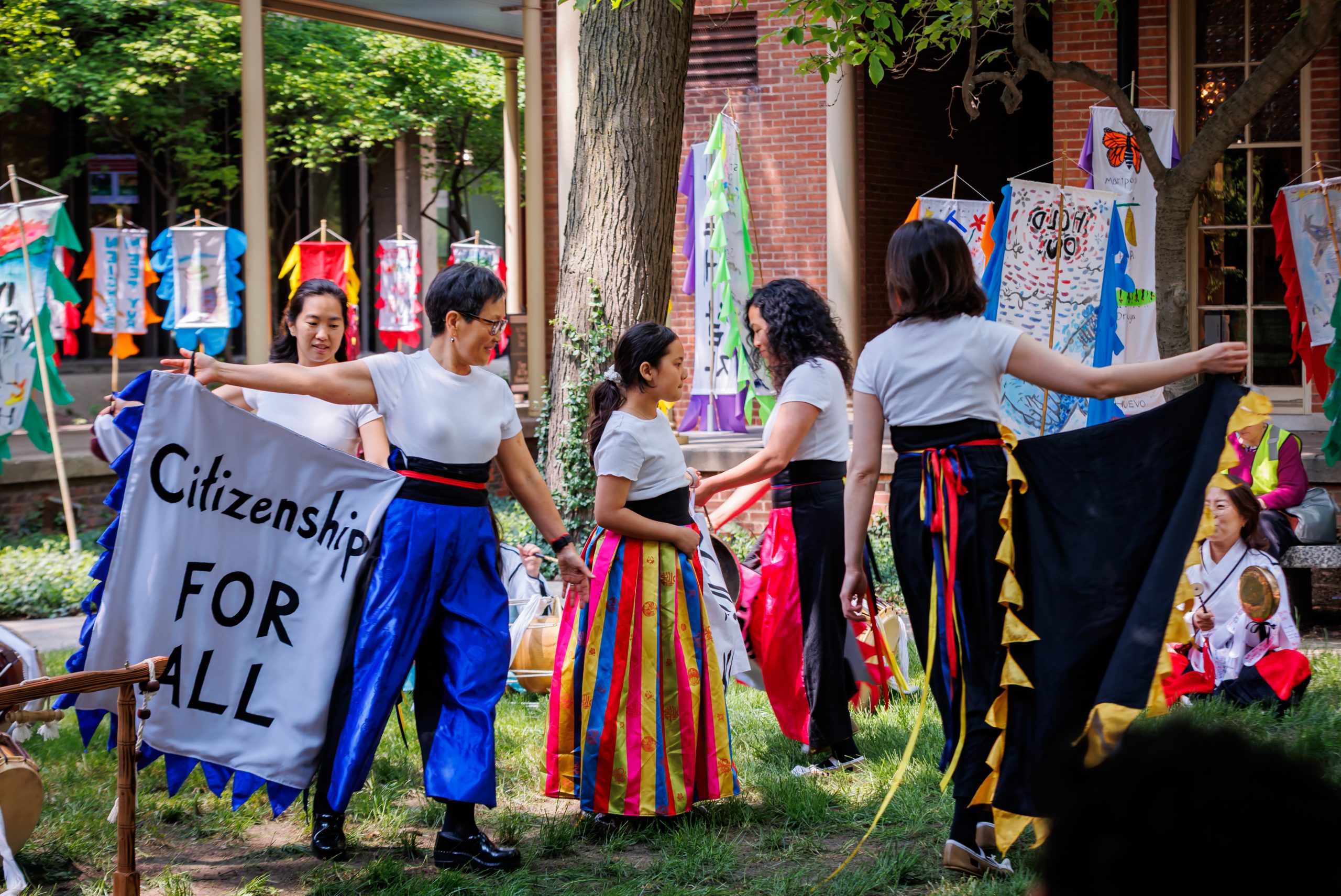A group of people in colorful, traditional costumes hold banners and gather outdoors. One banner reads “Citizenship FOR ALL.” Decorative flags and artwork hang in the background.