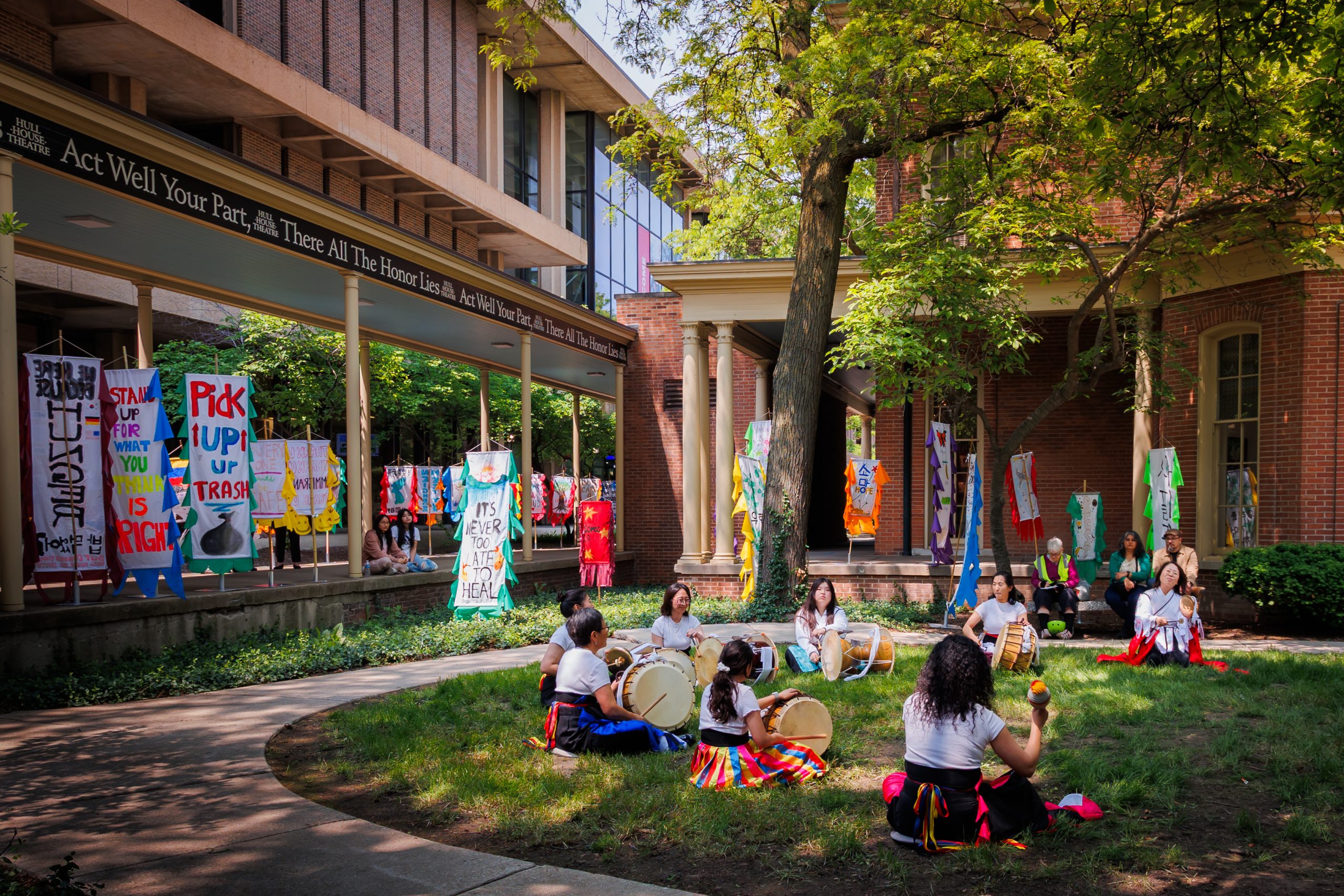 A group of people in colorful clothing play drums and shakers in a courtyard surrounded by trees, banners with messages hang around, and spectators watch from nearby. The setting appears to be a university or campus.